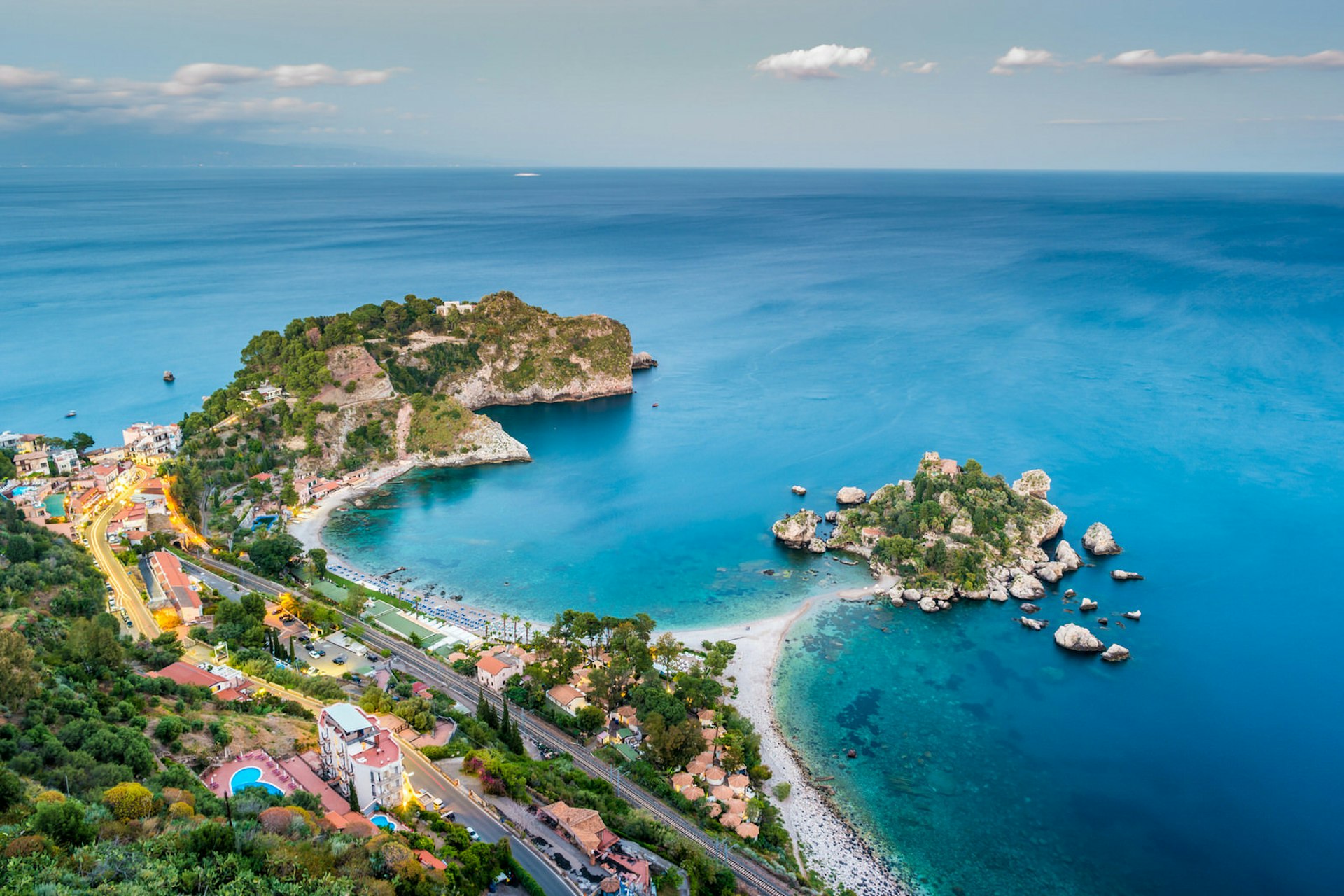 An aerial view of Isola Bella beach - a thin strip of white sand connecting the mainland to a small rocky island surrounded by shallow turquoise water.
