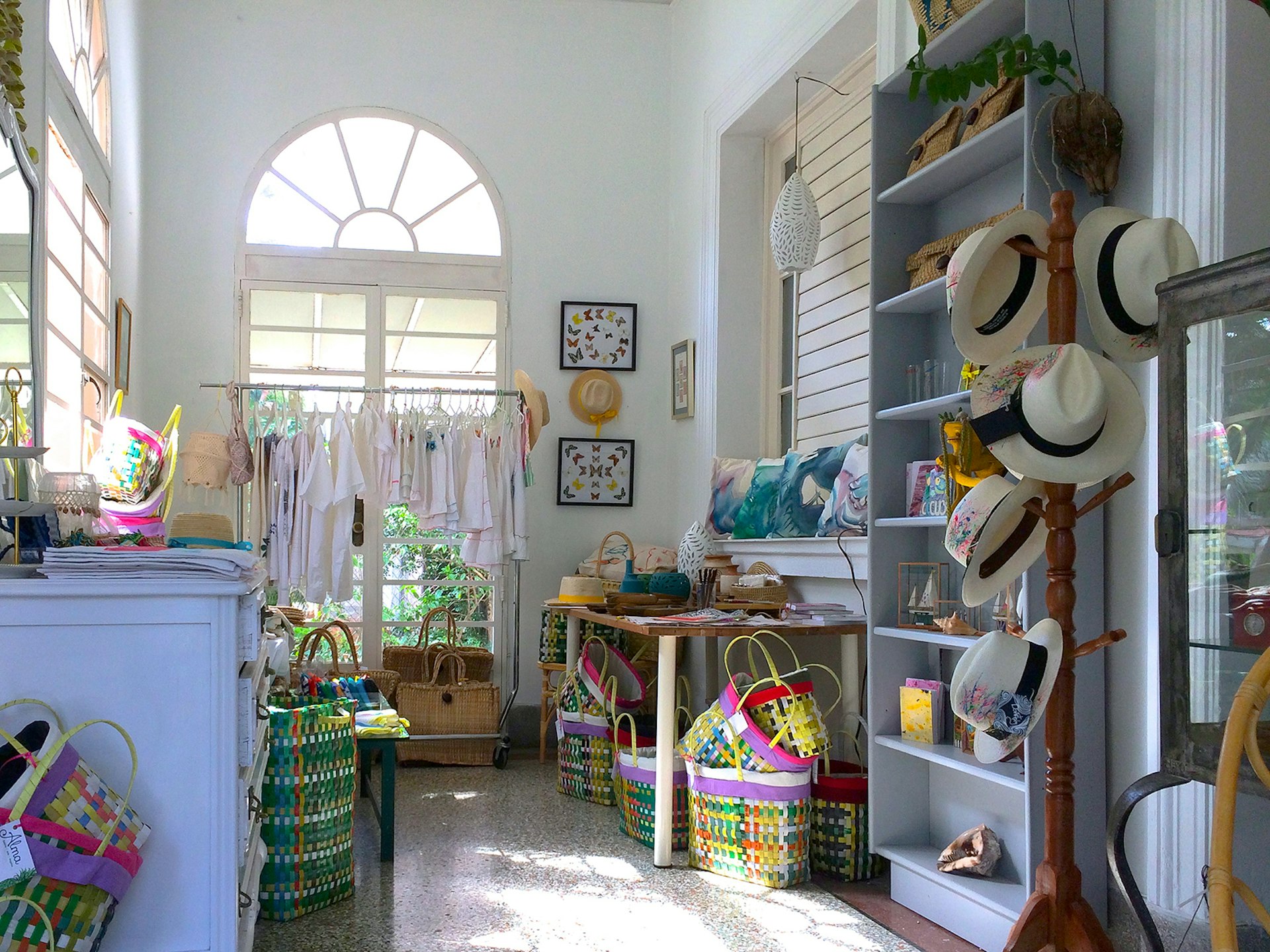 The stark white interior of the Alma boutique is filled with clothes hanging up near a window, a hat rack filled with Panama hats, a table filled with bags and hats; shopping choices abound in Havana 