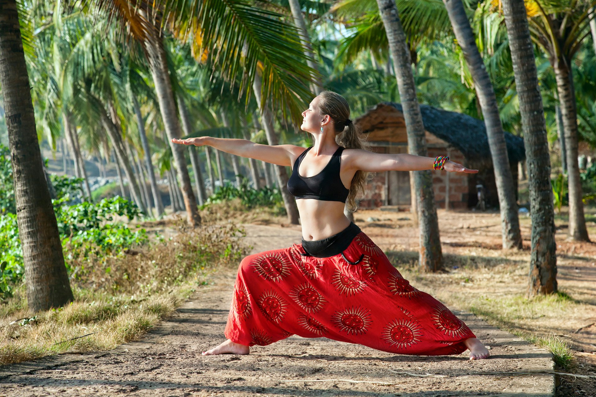 Yoga virabhadrasana II warrior pose by beautiful Caucasian woman in red Indian trousers with symbol om on the road in palm tree forest with house at background in India, Kerala, Varkala