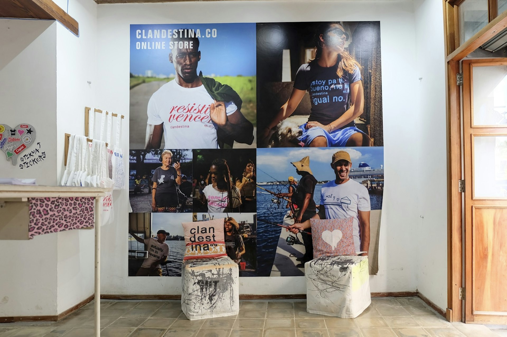 A large wall poster shows models wearing Clandestina t-shirts. In front of the poster are a pair of cushions with pillows on top. A row of canvas bags hang on a wall to the left. Shopping in Havana has vastly changed over the years