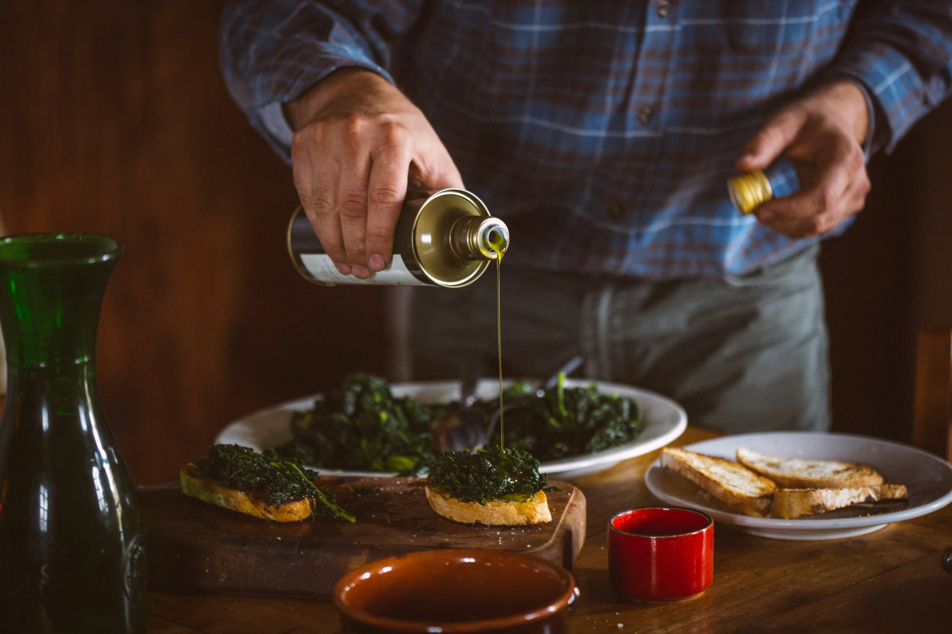 A close-frame image of a person pouring olive oil into a dish, wearing an apron. The image is cropped so we can just see their hands and torso standing behind a table laden with Italian oil, wines, and bruschetta. 