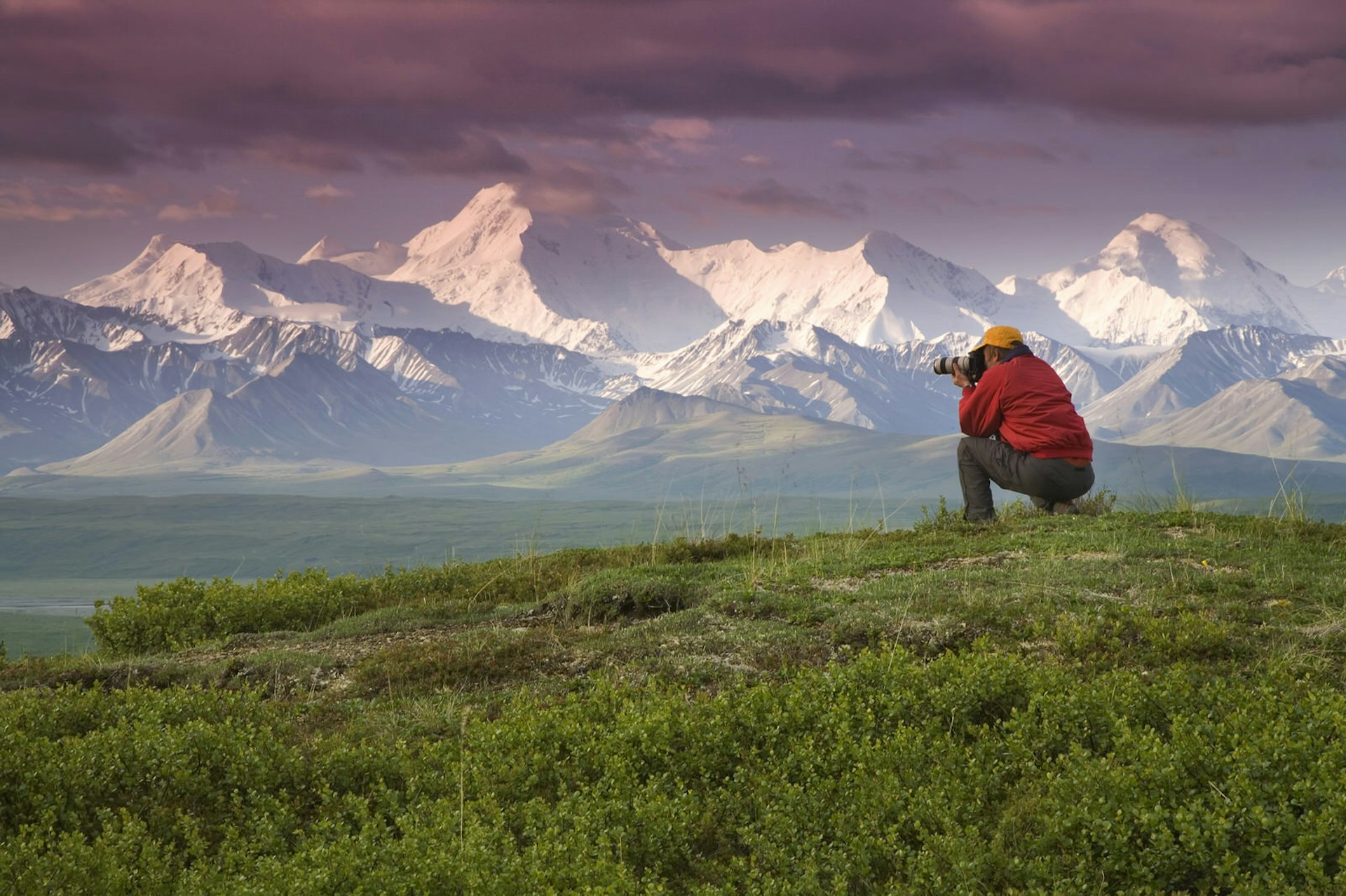 A man squats on a green hill taking photos of ice capped mountains in the distance.