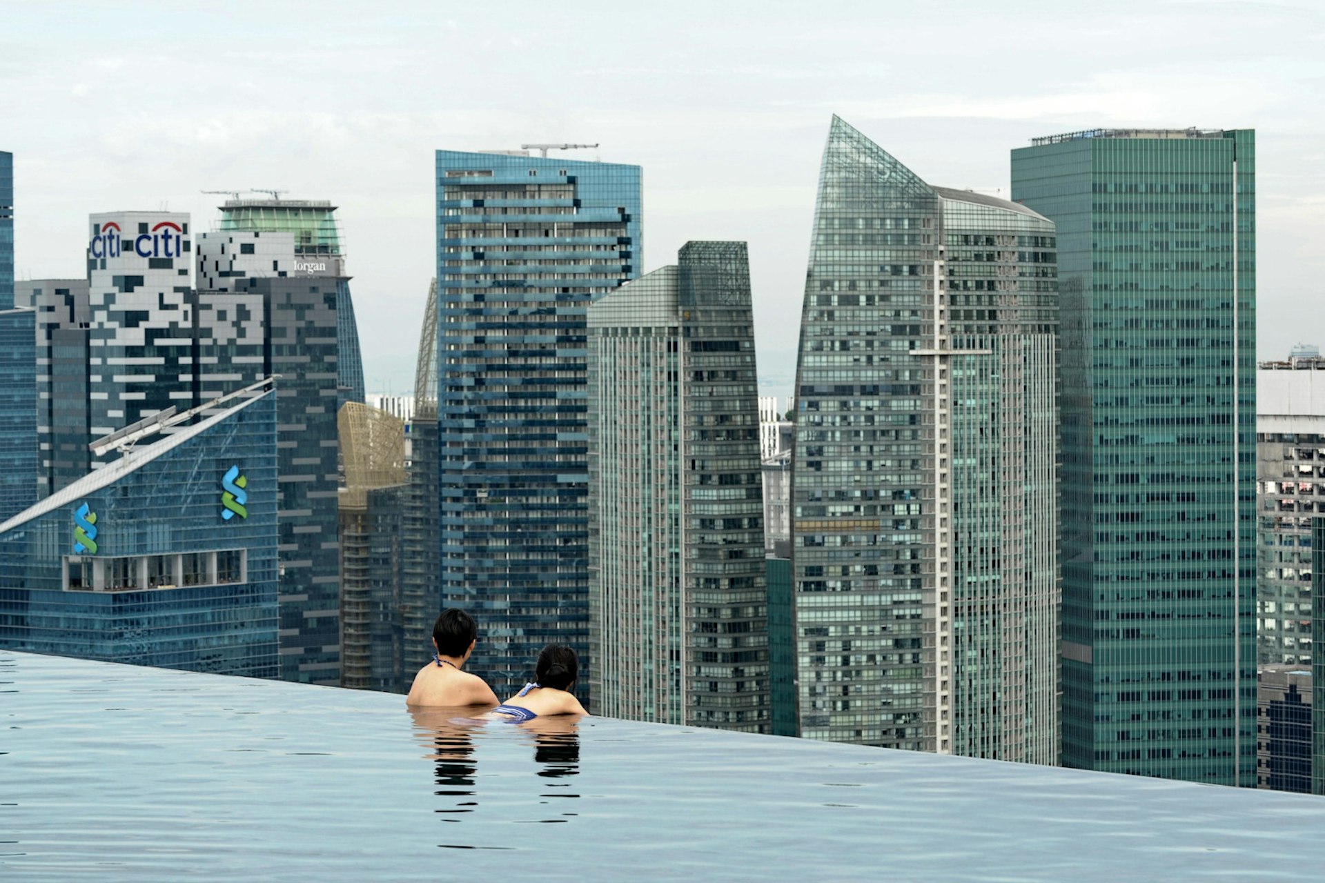 A couple in a rooftop pool looking out over a city skyscraper skyline.