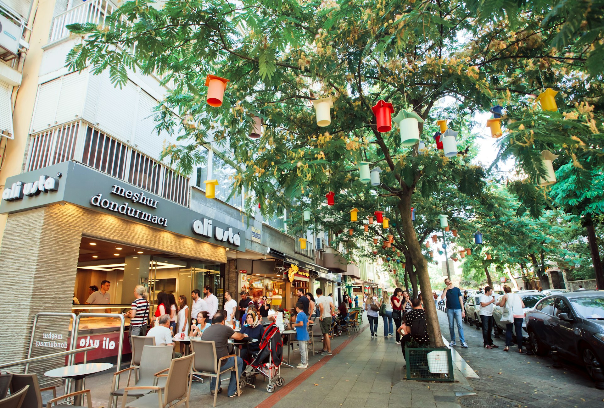 A tree-lined street with many people passing by. Some are sat outside a cafe to the left. The tree has colourful lanterns hanging from the branches