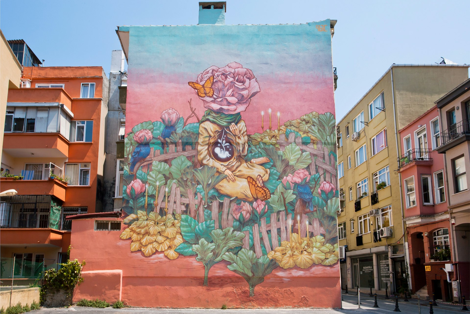 The side of a building is covered in a massive mural depicting a figure surrounded by plants. Instead of a face, the figure has a pink rose