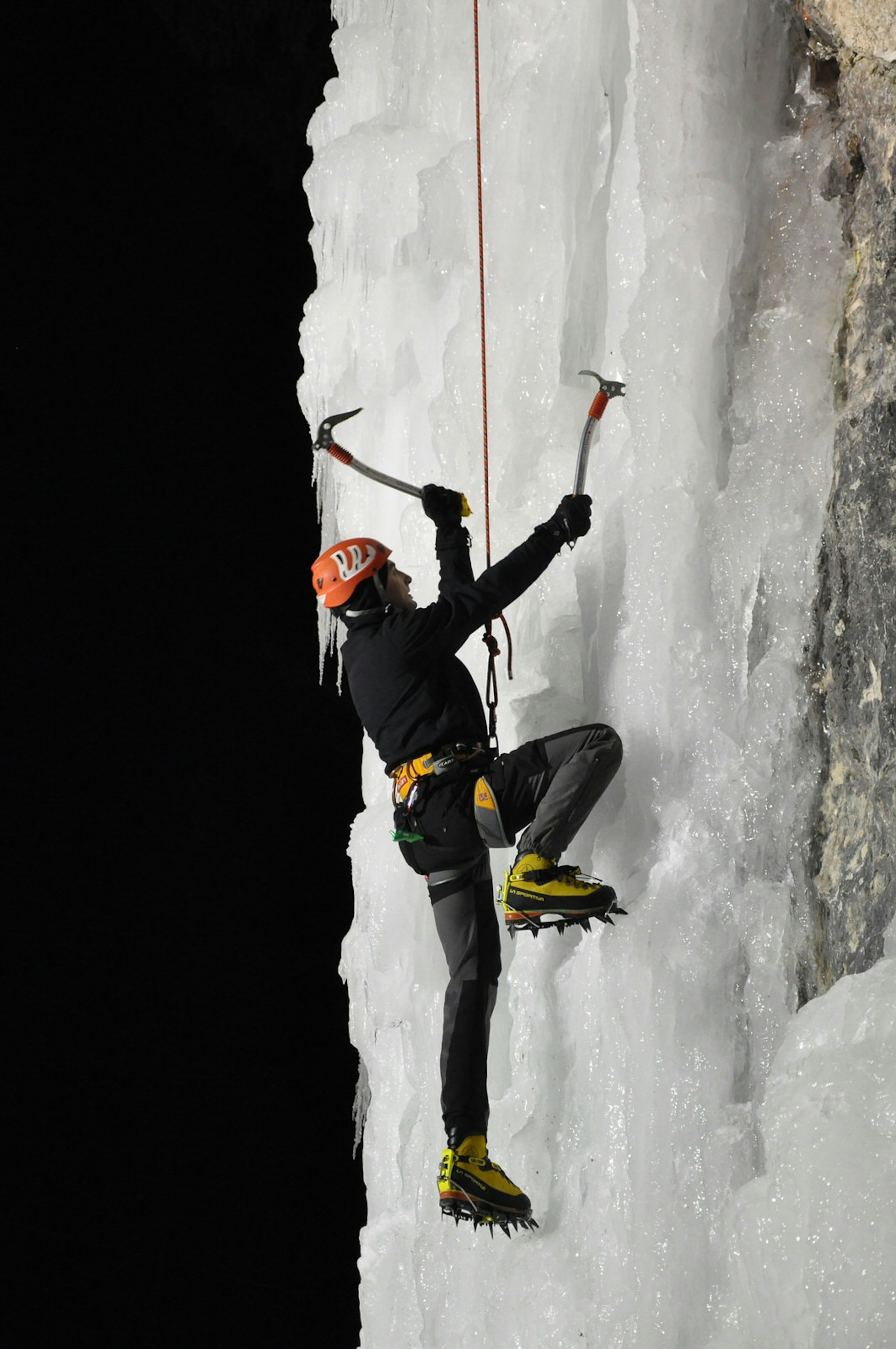 A person dressed in black wearing a safety harness, helmet and metal crampon uses two ice axes to scale a frozen wall of ice