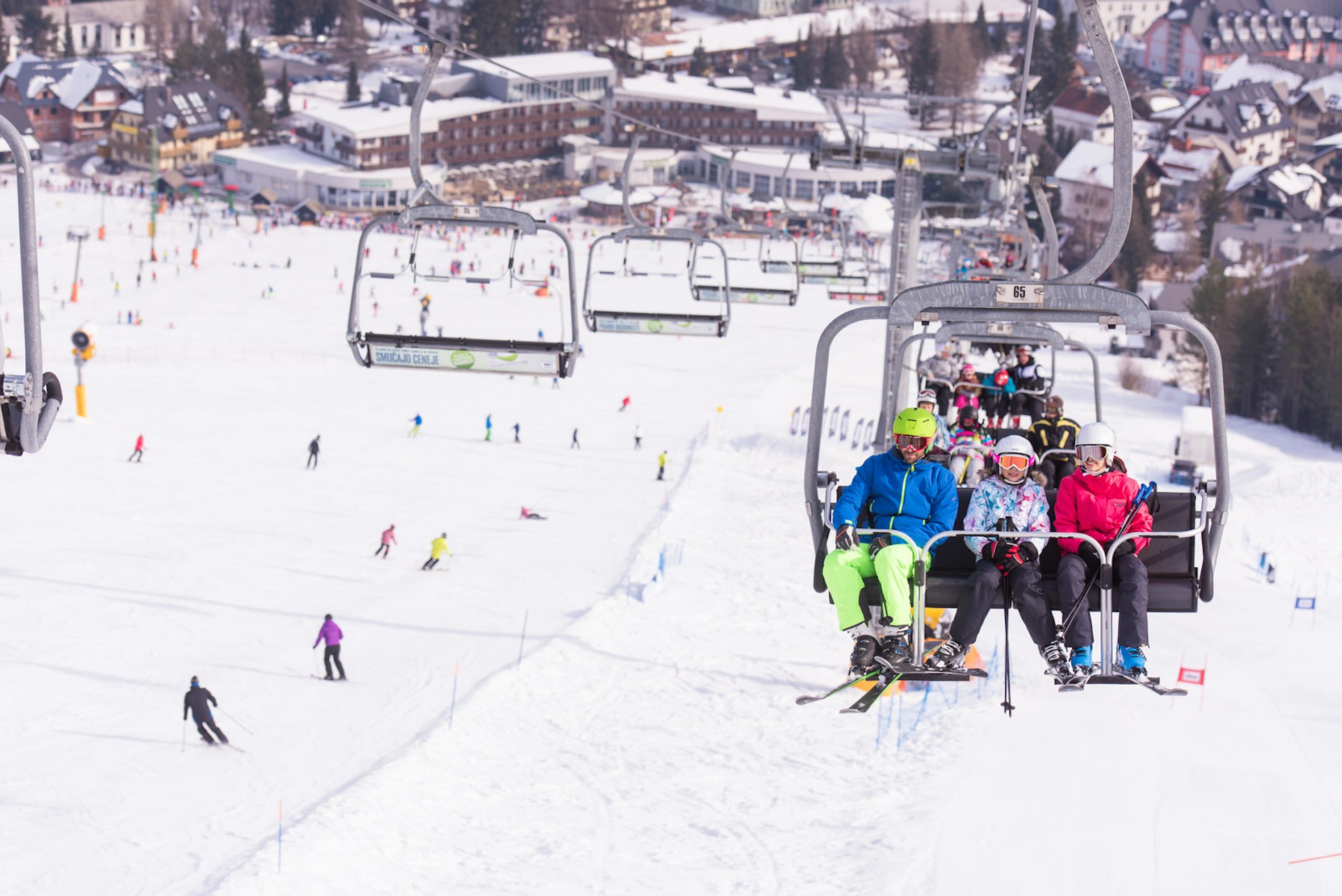 Ski lifts loaded with skiers in colourful gear go up the mountain, with empty chairs heading back down. Skiers can be seen on the slopes below