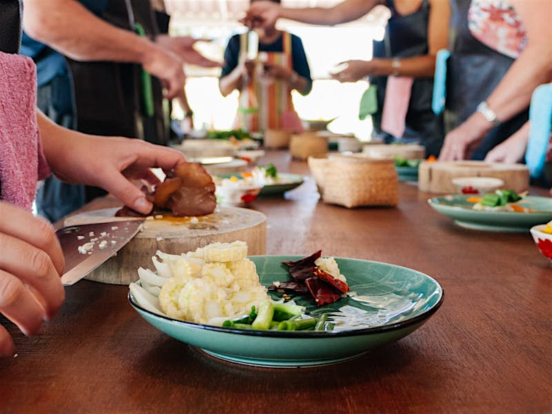 People stand either side of a table, which has a chopping board, ingredients and plates of Thai food
