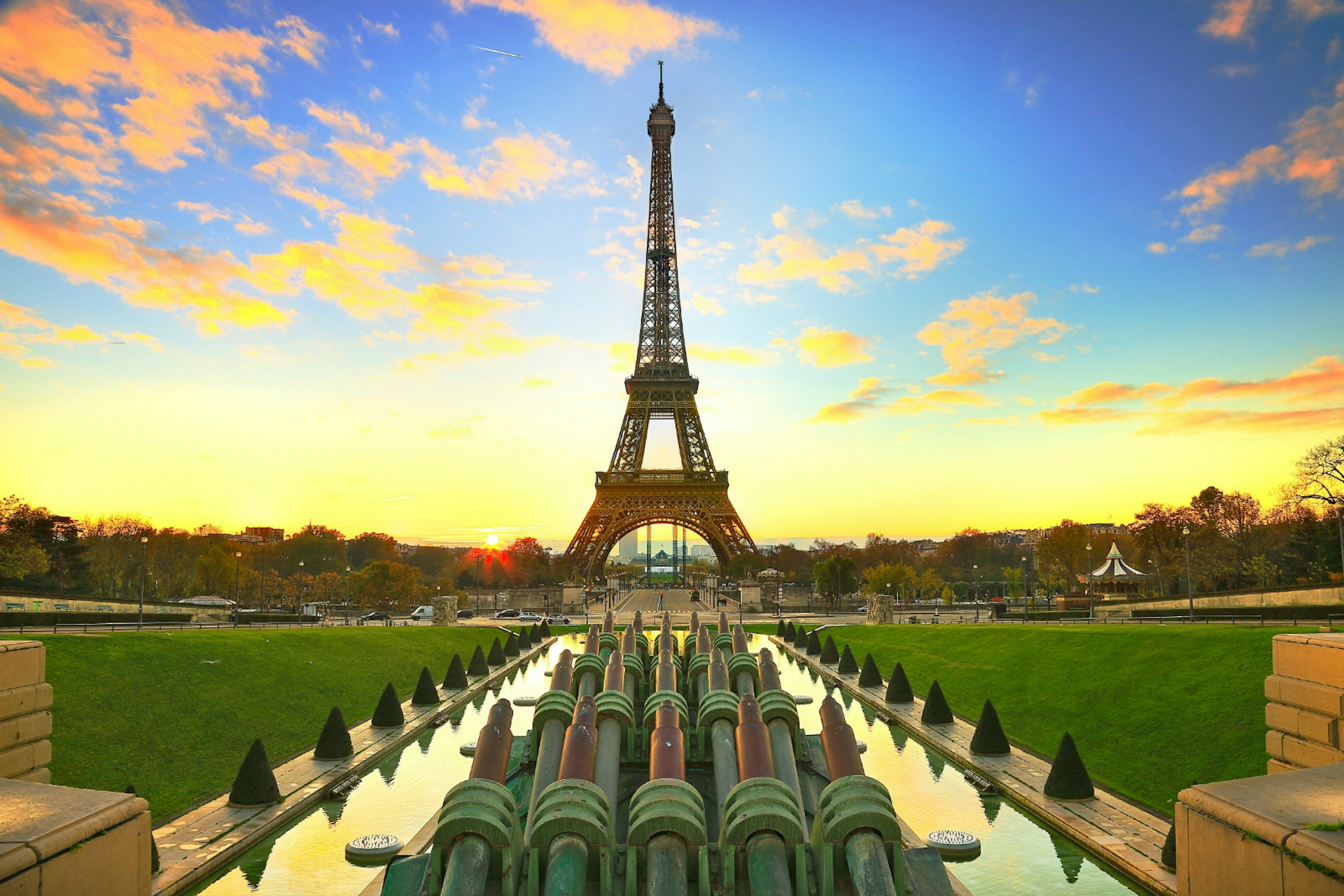 The sun rises behind the Eiffel Tower, turning the sky yellow, blue and orange. The photo is taken from behind a sculpture that is flanked by water and green lawns