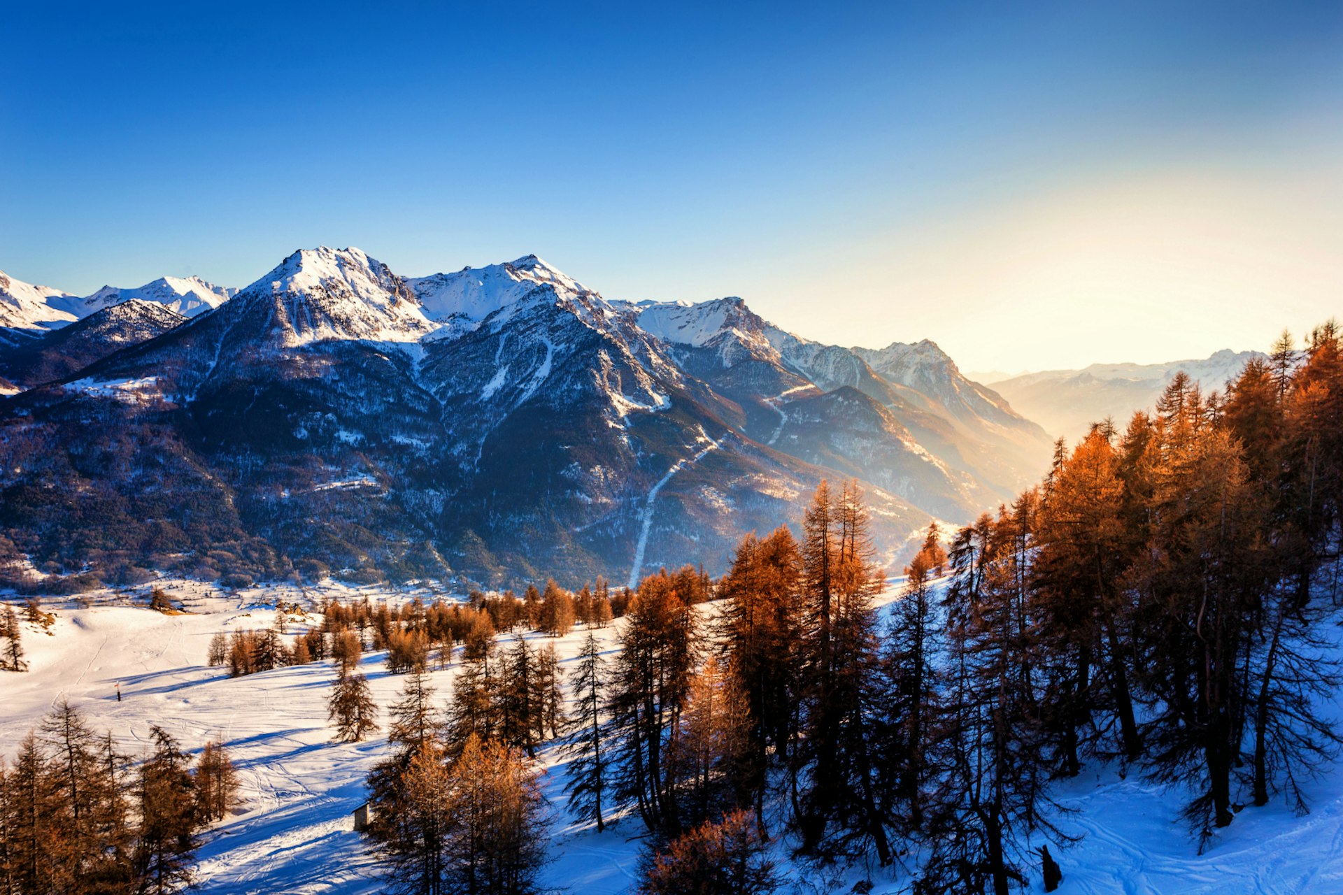The sun sets over Briançon in the French Alps. There are sparse coniferous trees in the foreground, sitting on a blanket of snow, with rugged mountain peaks in the background, criss-crossed with trails