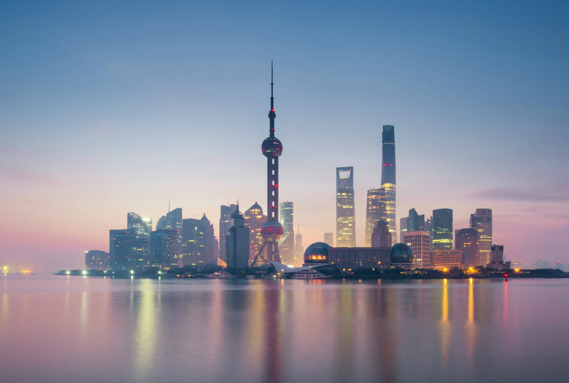 The skyline of Pudong in Shanghai, surrounded by pink water and pale pink and blue light in the sky as the sun sets