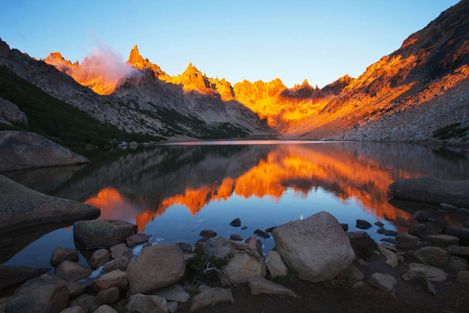 The irresistible mountain lake at Refugio Frey in the Argentine Andes at sunset. The still blue water reflects the jagged orange peaks that are bathed in the suns warm light. 