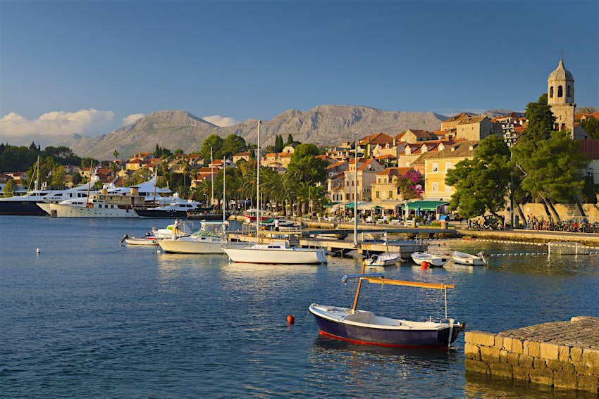 Cavtat's pretty marina under late afternoon sun: boats are moored on the water, with a stone church spire, orange-roofed buildings and trees along the shore; barren mountain peaks form a dramatic backdrop. 