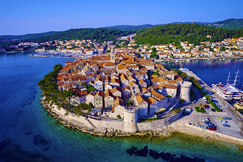 An aerial view of Korčula's beautiful walled old town, which resembles a citadel jutting out into stunning turquoise water; round defensive towers punctuate the wall, which protects the orange-roofed buildings of the town. Beyond are a harbour and the green hills of the island's interior. 