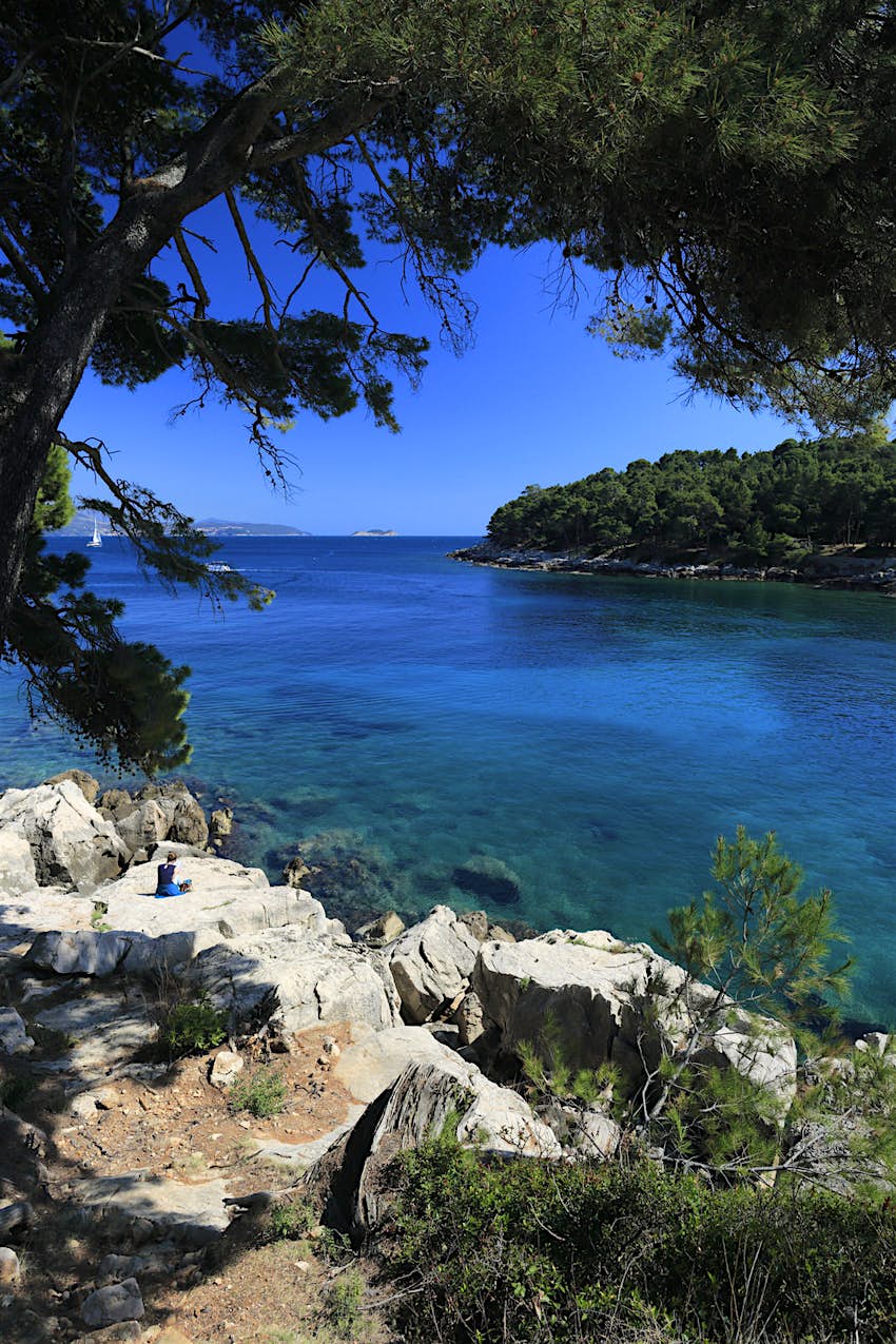 A lone female traveller wearing a blue skirt and a navy top sits on a rock next to brilliant blue water off Lokrum Island; the scene is shaded by pine trees and another pine-clad island is visible across the water.