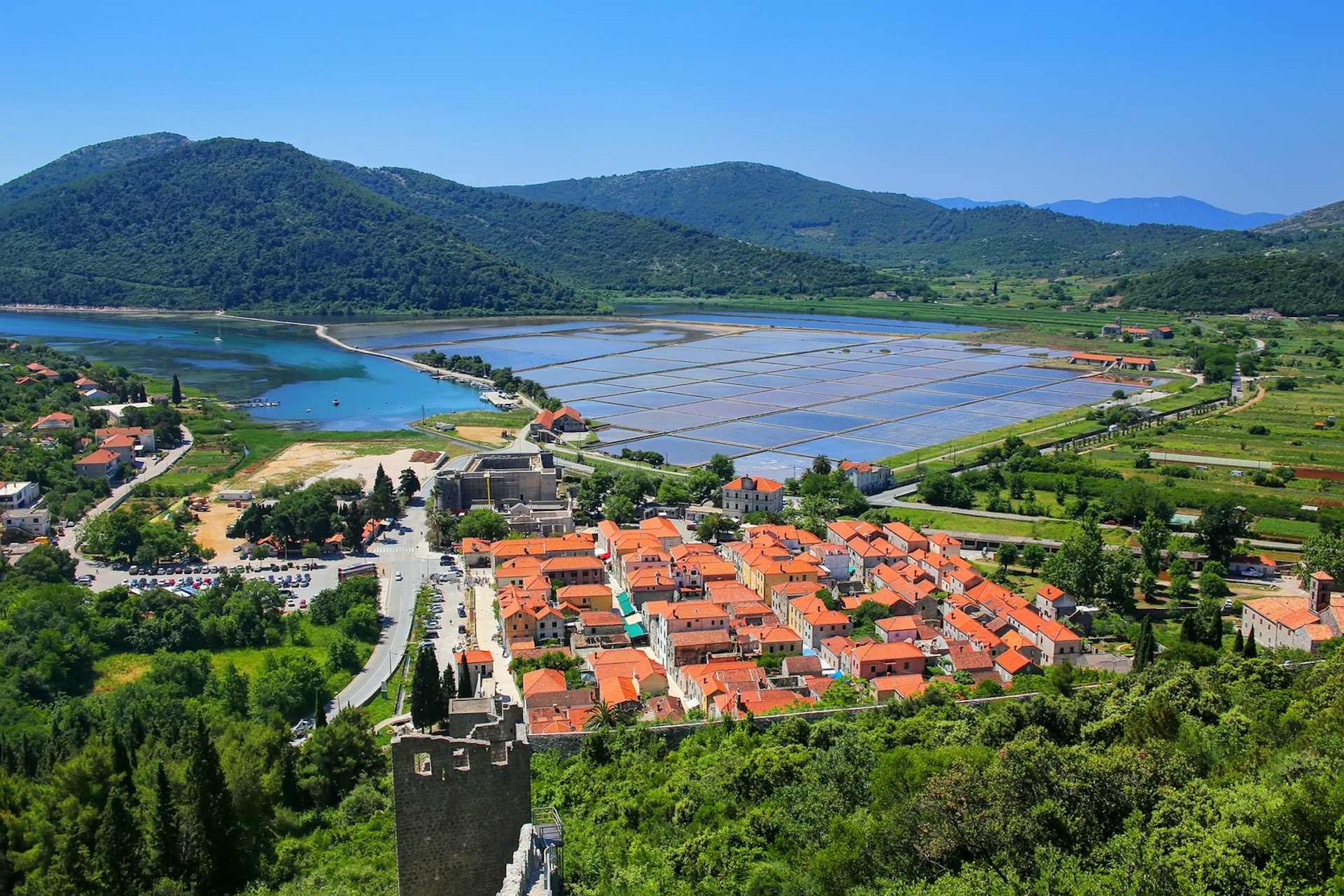 View of Ston town and salt pans from above; the town is a compact square of orange roofs, surrounded by green fields and trees; beyond are salt pans and forested peaks.