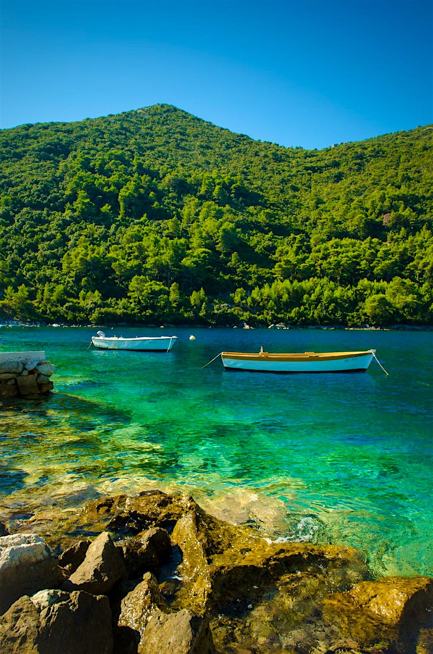 Two boats sit moored in the idyllic turquoise water of Mljet National Park; there are rocks in the foreground and forest-clad hills on the other side of the water.