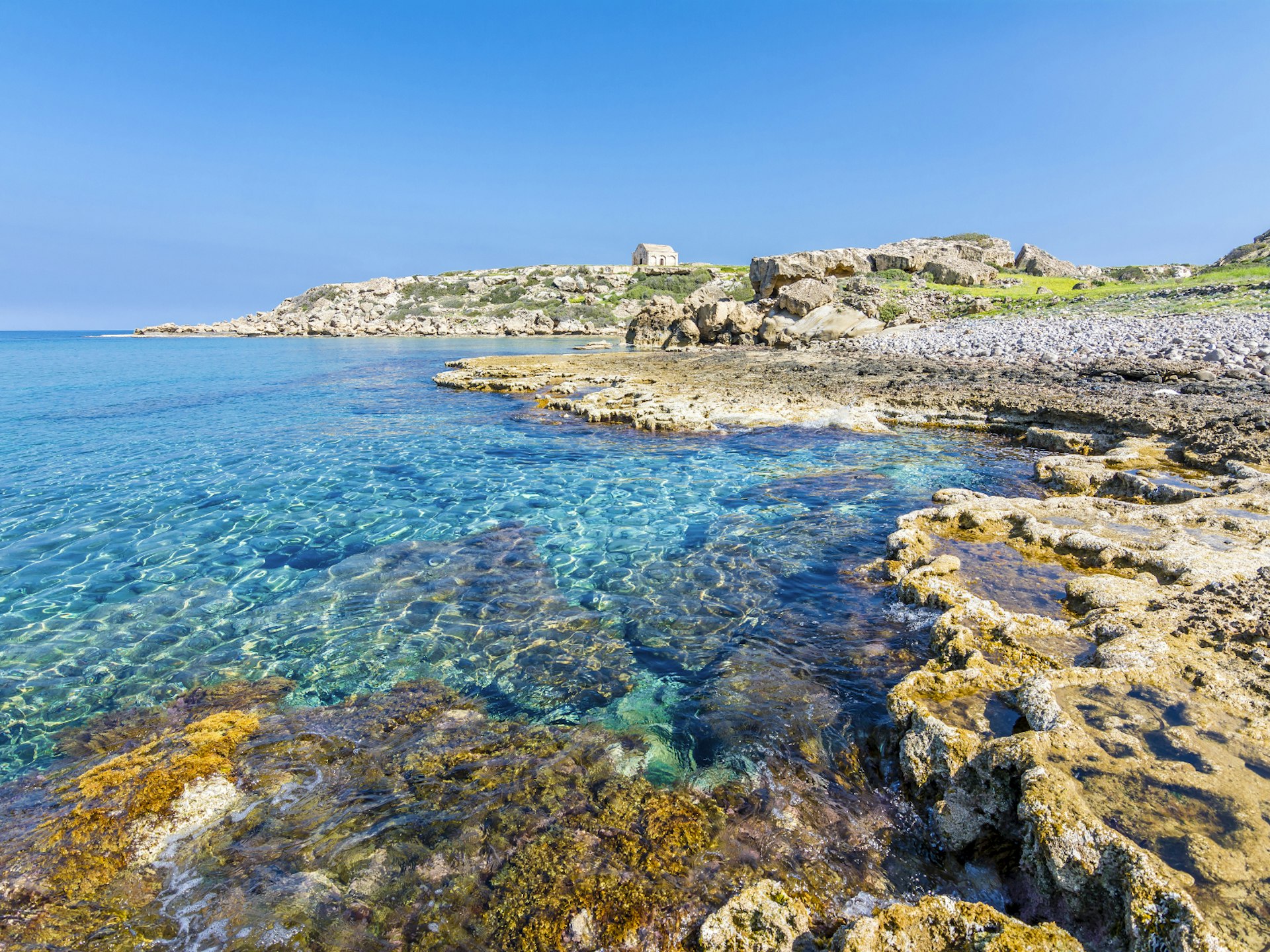 A rocky shoreline with a small stone building in the distance. The sea is clear and very still.