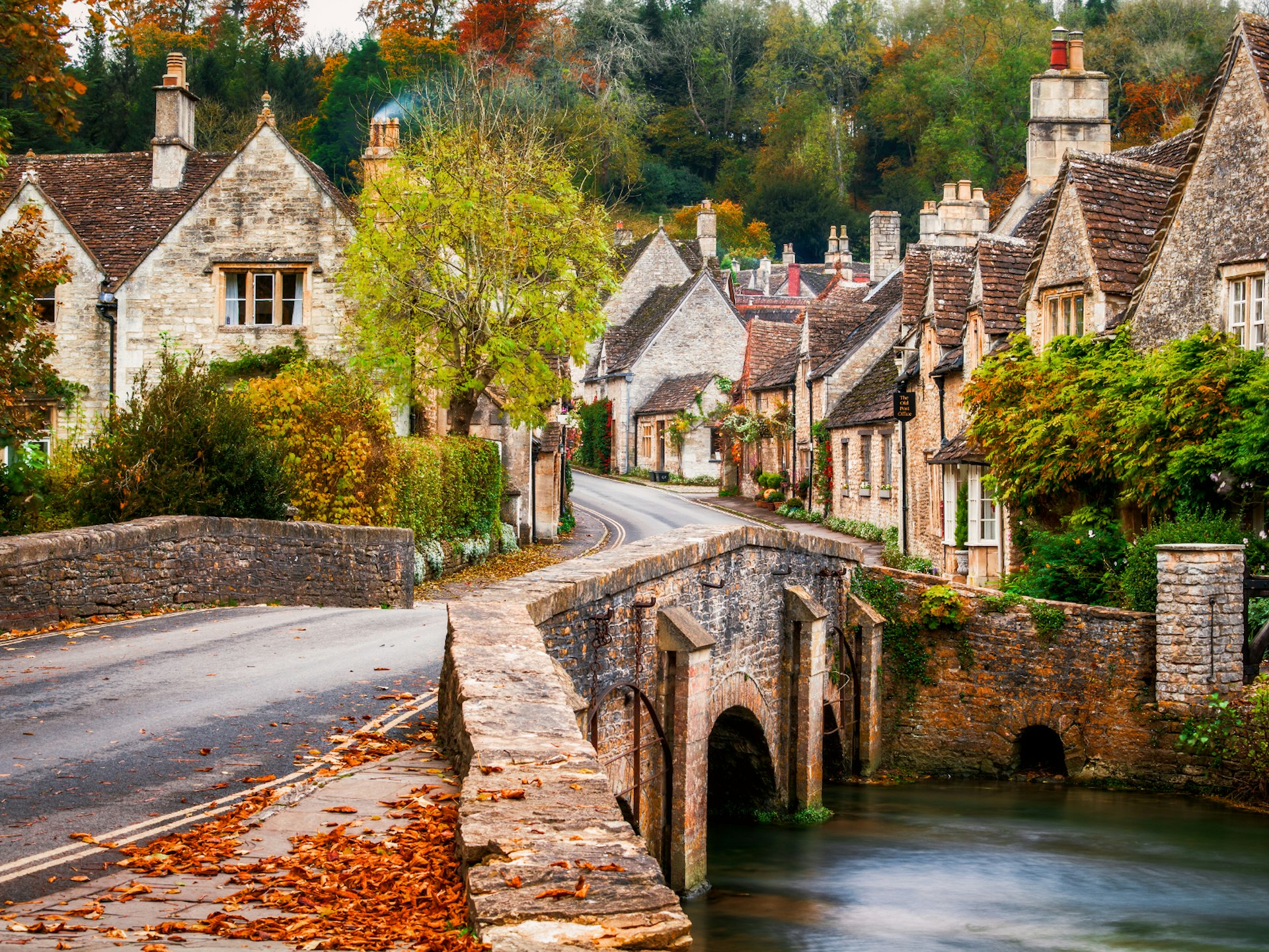 A road over a stone bridge leads to traditional cottages with pointed roofs and chimneys with autumn foliage all around