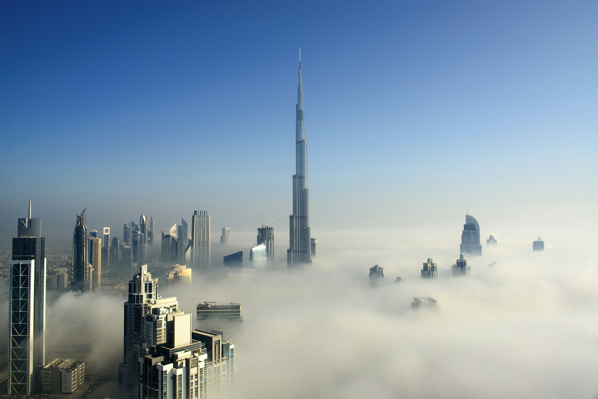 Foggy morning in downtown Dubai, United Arab Emirates. The city's many skyscrapers are poking through thick cloud, into a blue sky, with the Burj Khalifa in the centre of the frame.