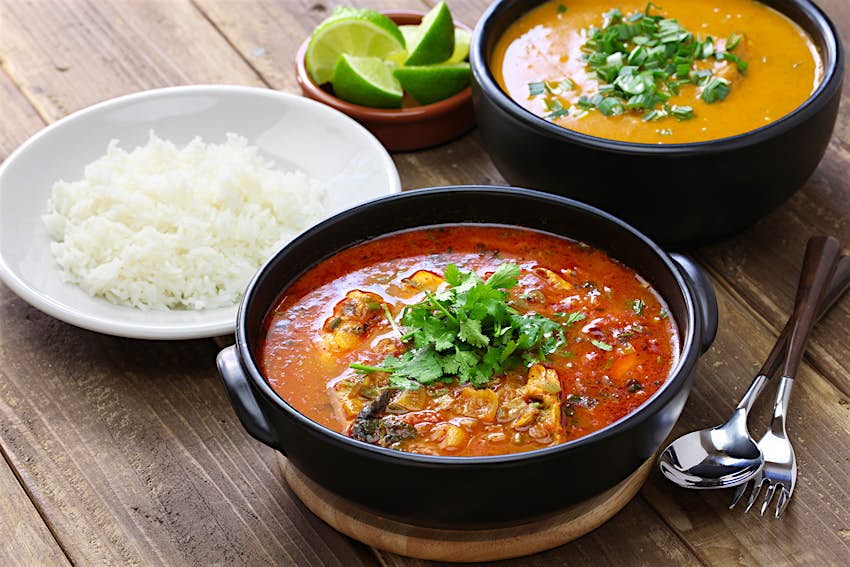 A bowl of red stew topped with cilantro sits on a wooden table alongside a plate of rice and a bowl of creamy orange soup. South America.