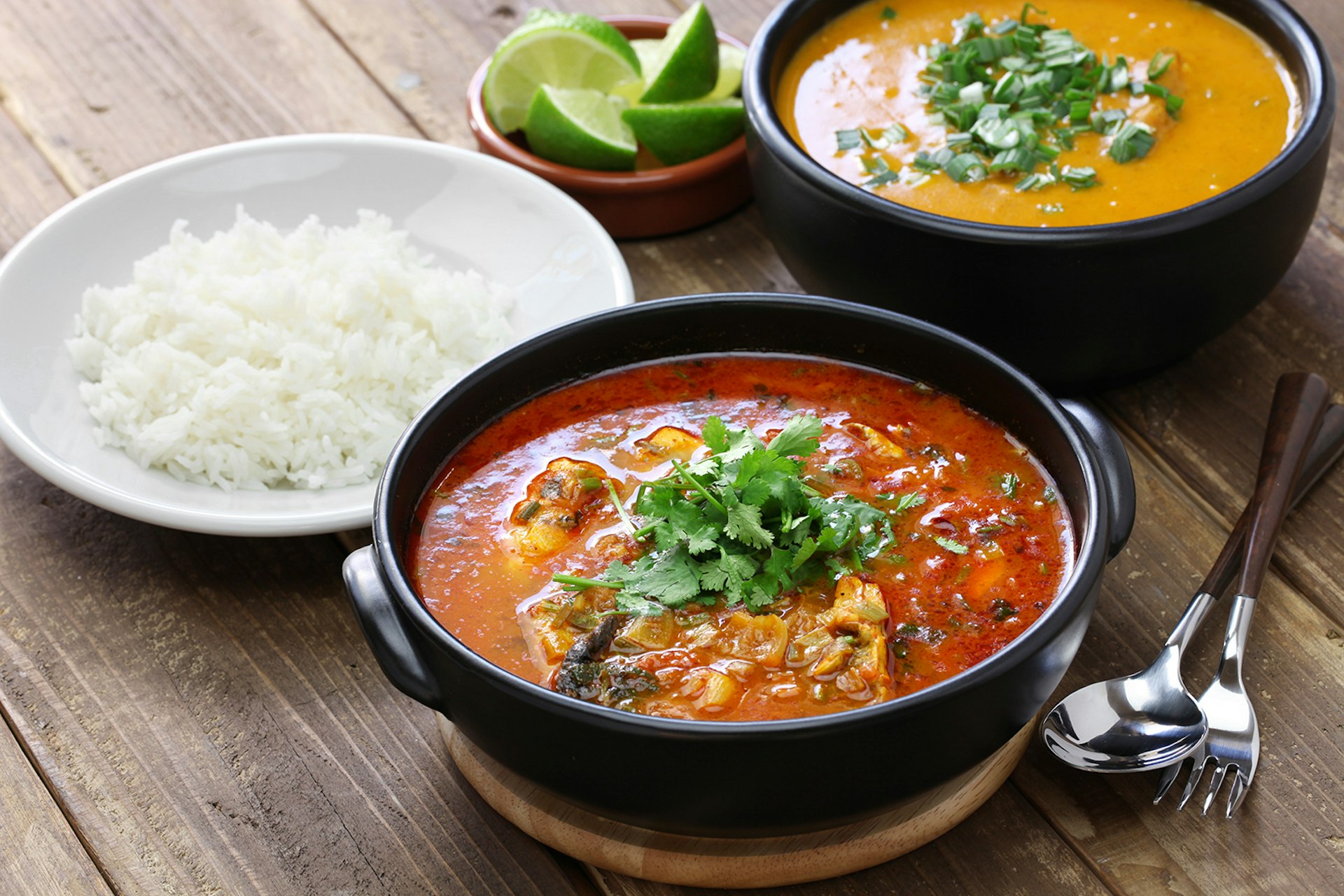A bowl of red stew topped with cilantro sits on a wooden table alongside a plate of rice and a bowl of creamy orange soup. South America.