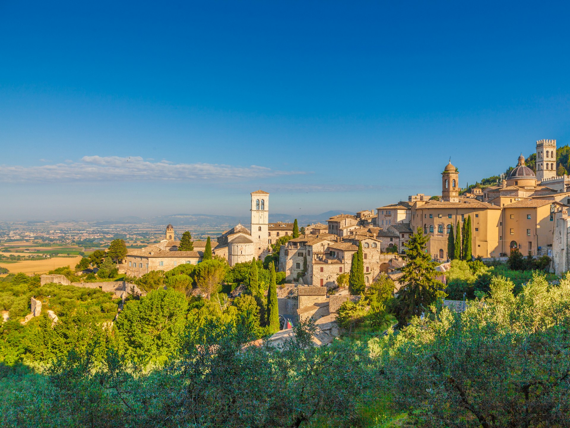 The sandstone-coloured buildings of Assisi glow in the sunshine, with a clear blue sky behind and countryside in the distance