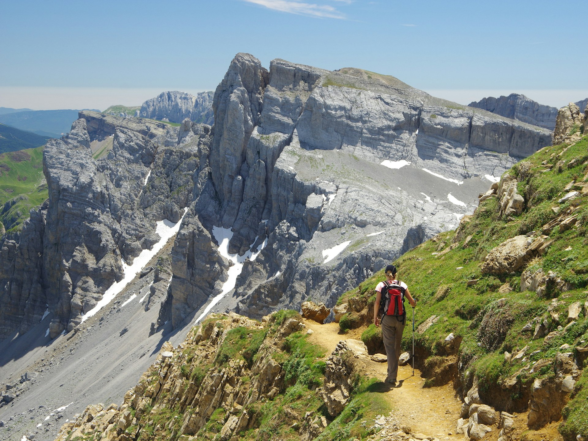 A female hiker walks a narrow mountain path with peaks ahead of her, on a sunny day in the Spanish Pyrenees