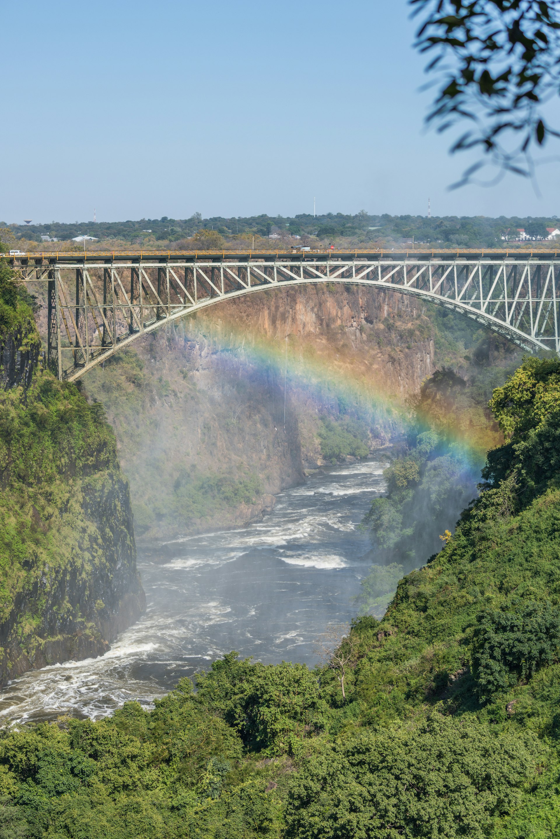 A misty rainbow is seen in front of the bridge, which is high above the Zambezi river. Two bungee jumpers can just be seen dangling below the bridge