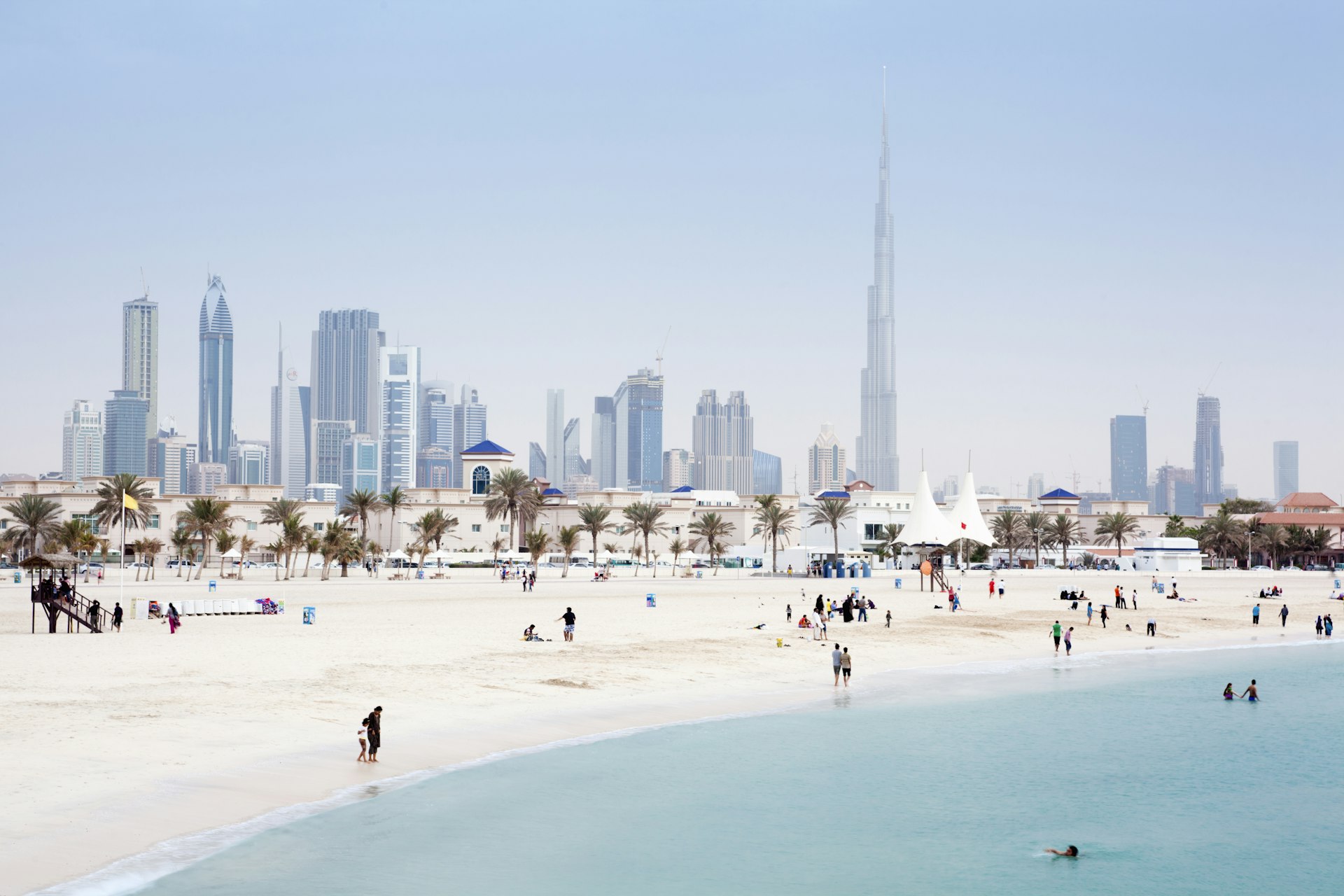 People swimming in the sea and walking along the shore of Jumeirah Beach with the Dubai skyline in the background