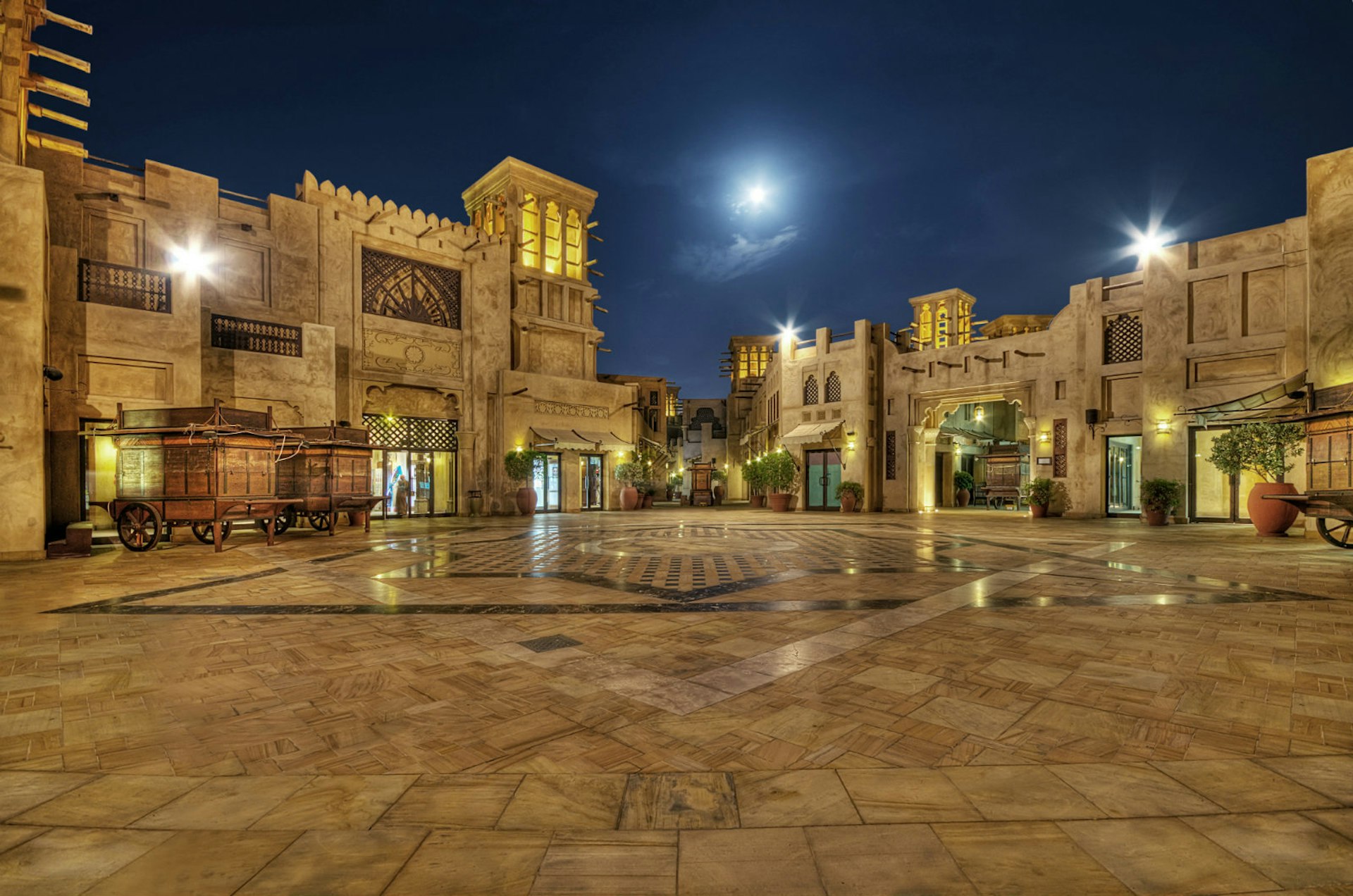 The moon rises over a darkened square in Dubai, United Arab Emirates. Th ground is elaborately tiled and there are wooden carts outside old-fashioned shop fronts. 