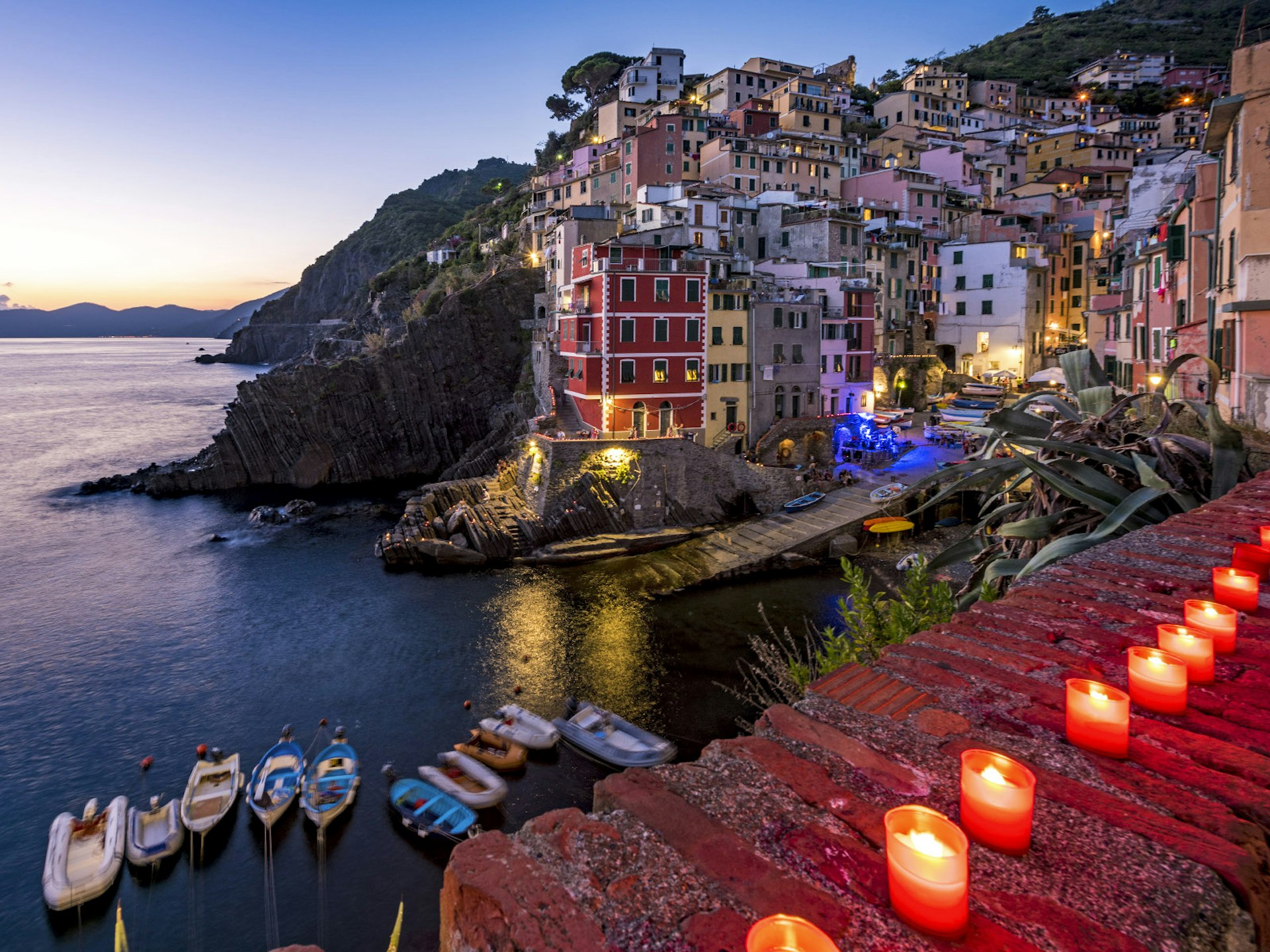 Riomaggiore, with brightly coloured buildings on the rocky hillside, and a handful of small boats docked in the harbour. Candles are lit along the wall in the foreground