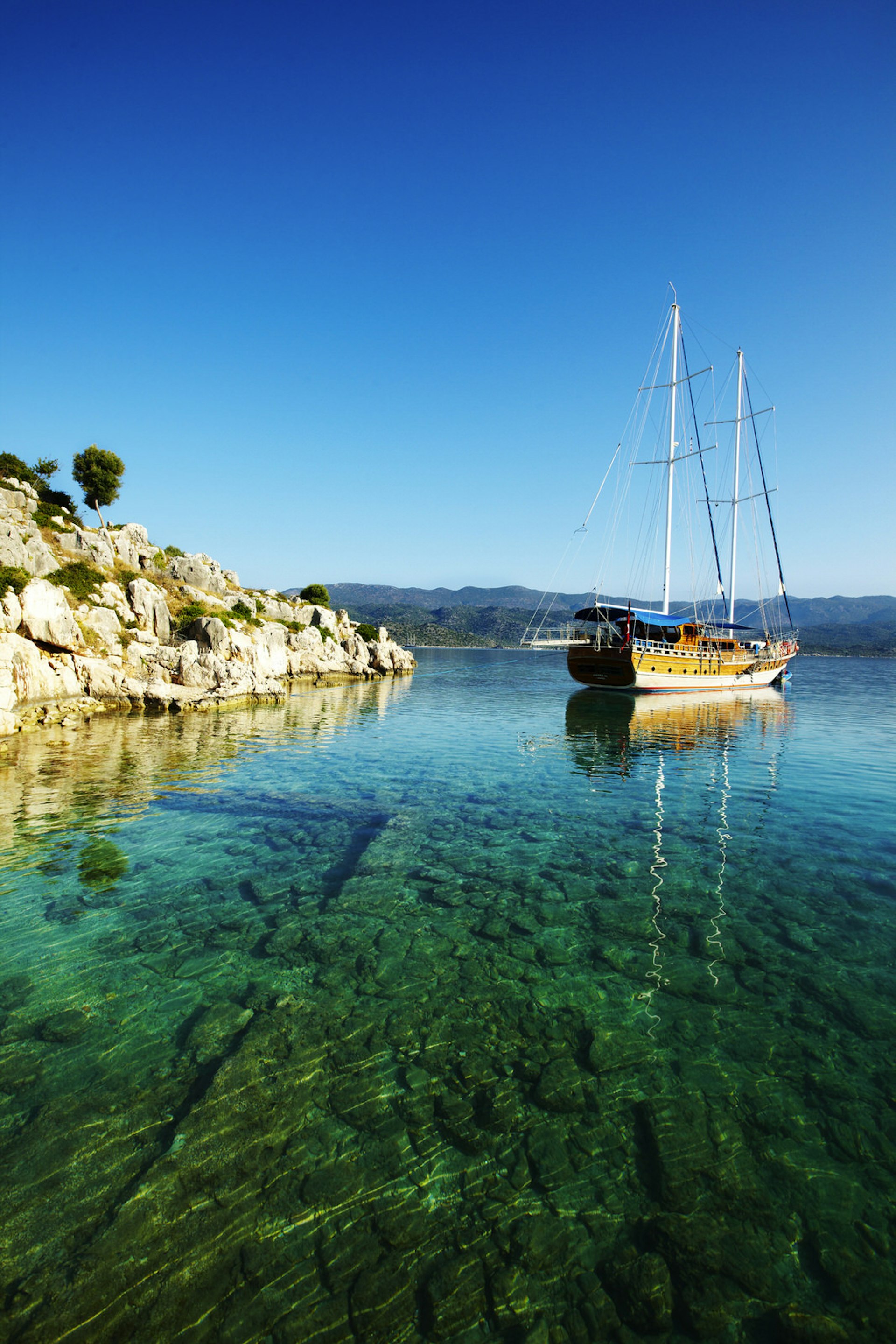 Sunken ruins can be seen below the surface of the clear blue-green waters, with a traditional wooden Turkish sailing boat in the background