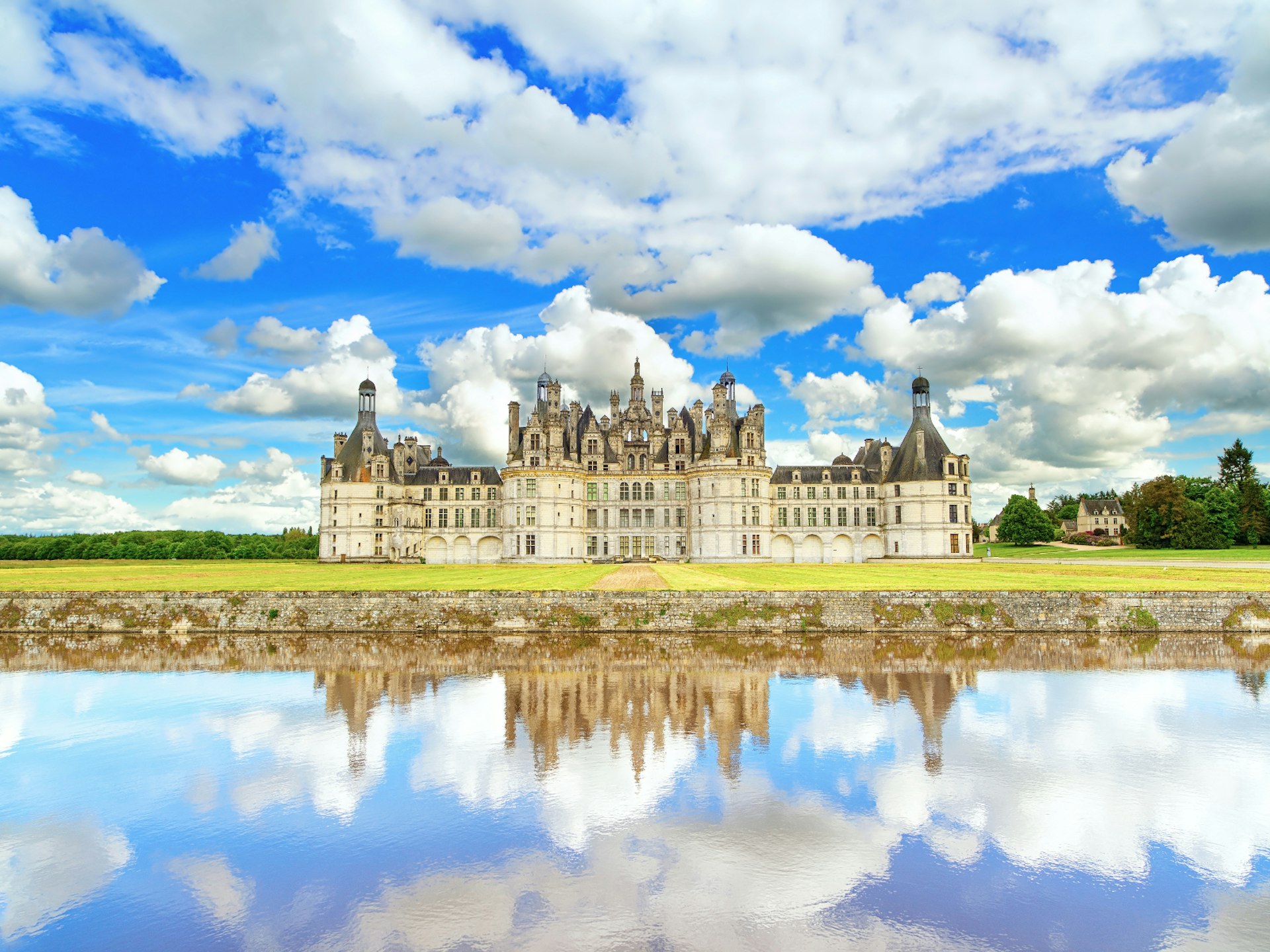 Chateau de Chambord, an impressive white-brick renaissance building with turrets and a grey slate roof. There is a large body of still water in front of the castle that perfectly mirrors the cloudy blue sky above
