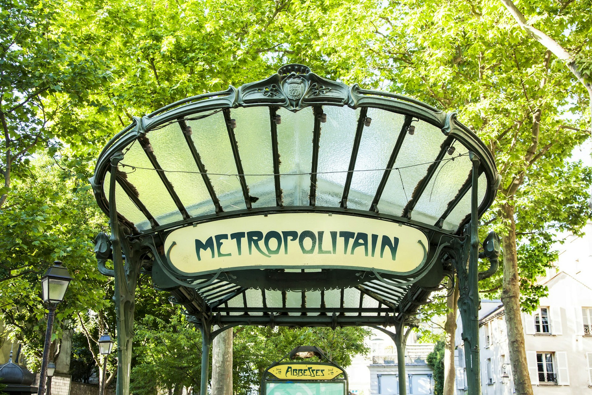 Art deco sign for the Abbesses metro station stating 'metropolitain' with green trees in the background