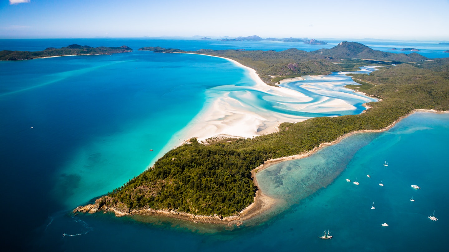 A small peninsula covered in vegetation in the foreground with the turquoise swirls of sea mixing with the white sands at Whitehaven Beach behind