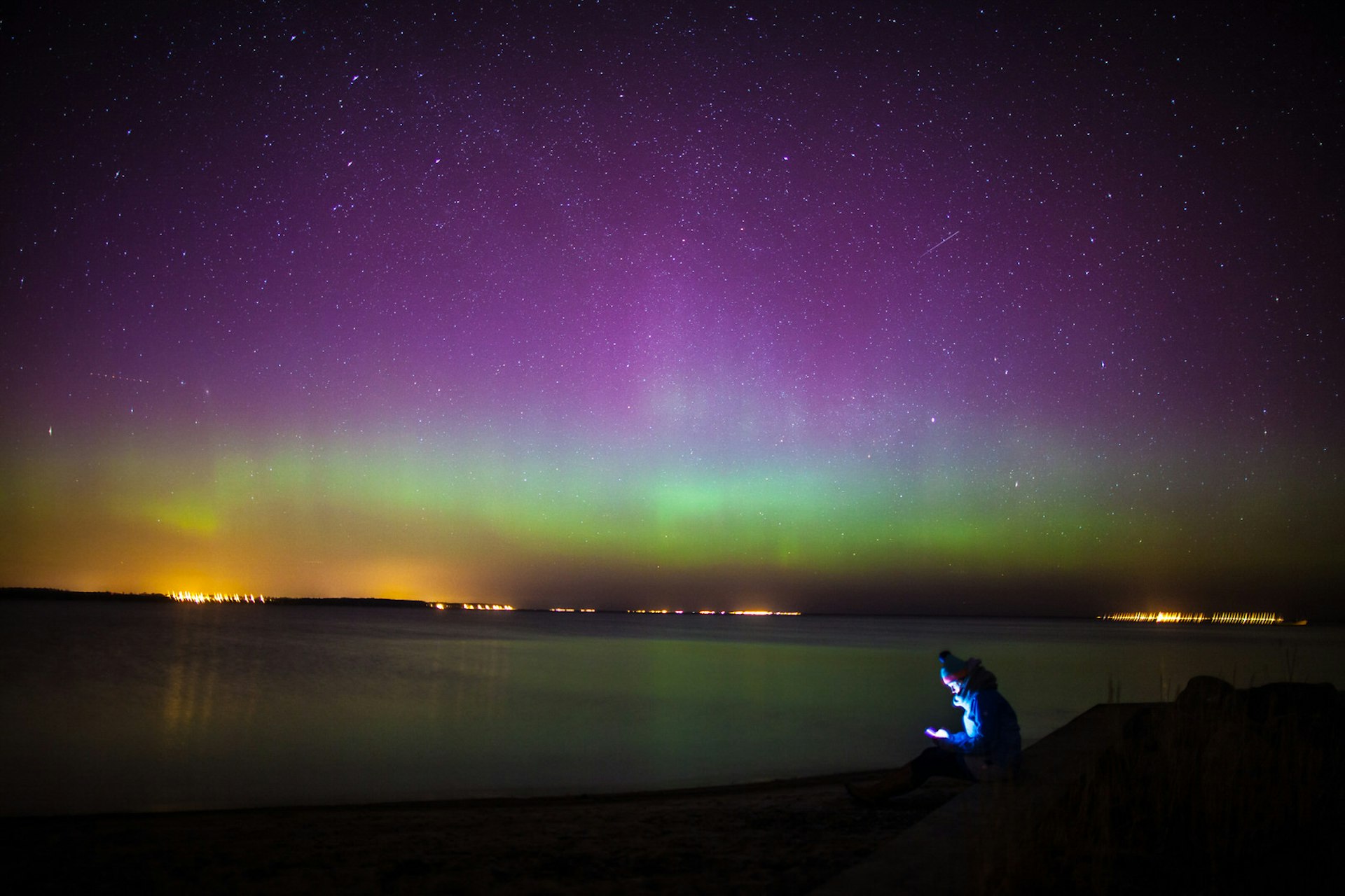 A figure sits huddled over a light source while the Northern Lights glow green and purple overhead