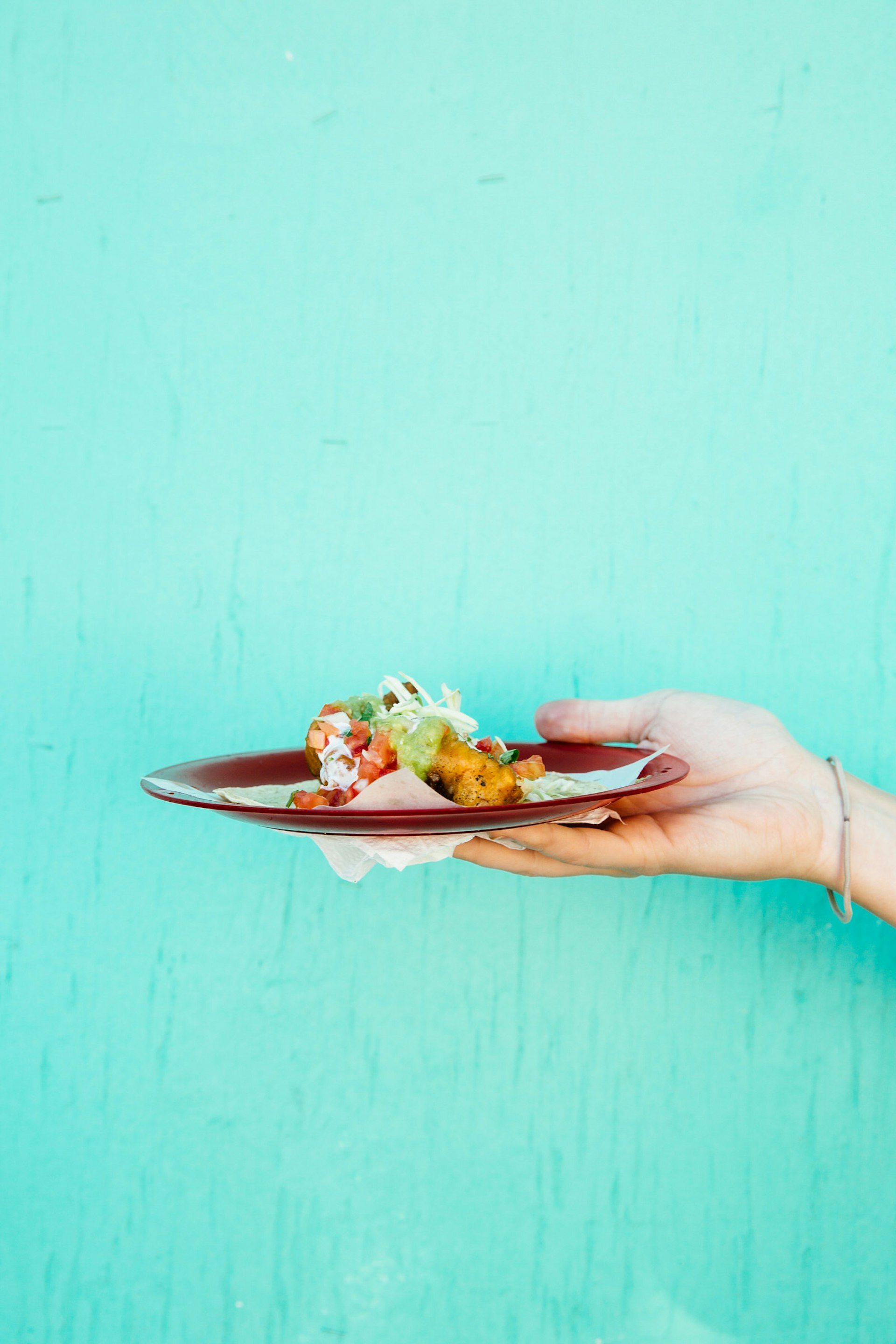 A hand holds a plate of fish tacos in front of a bright turquoise wall © Cultura Exclusive / Tanveer Badal / Getty Images
