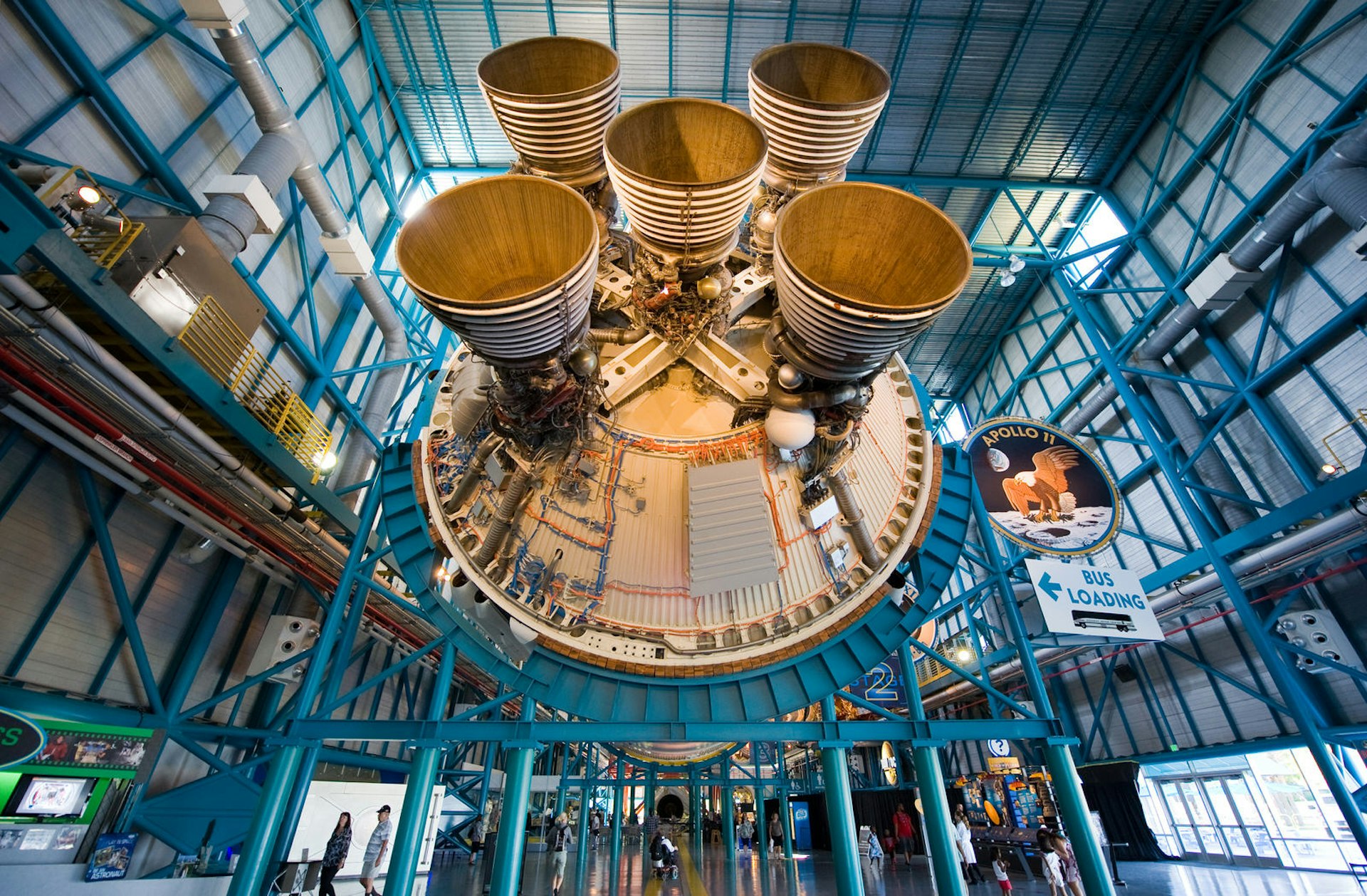 An upwards shot looking towards the five engines of the second stage of the Saturn 5 rocket. A few people mingle below