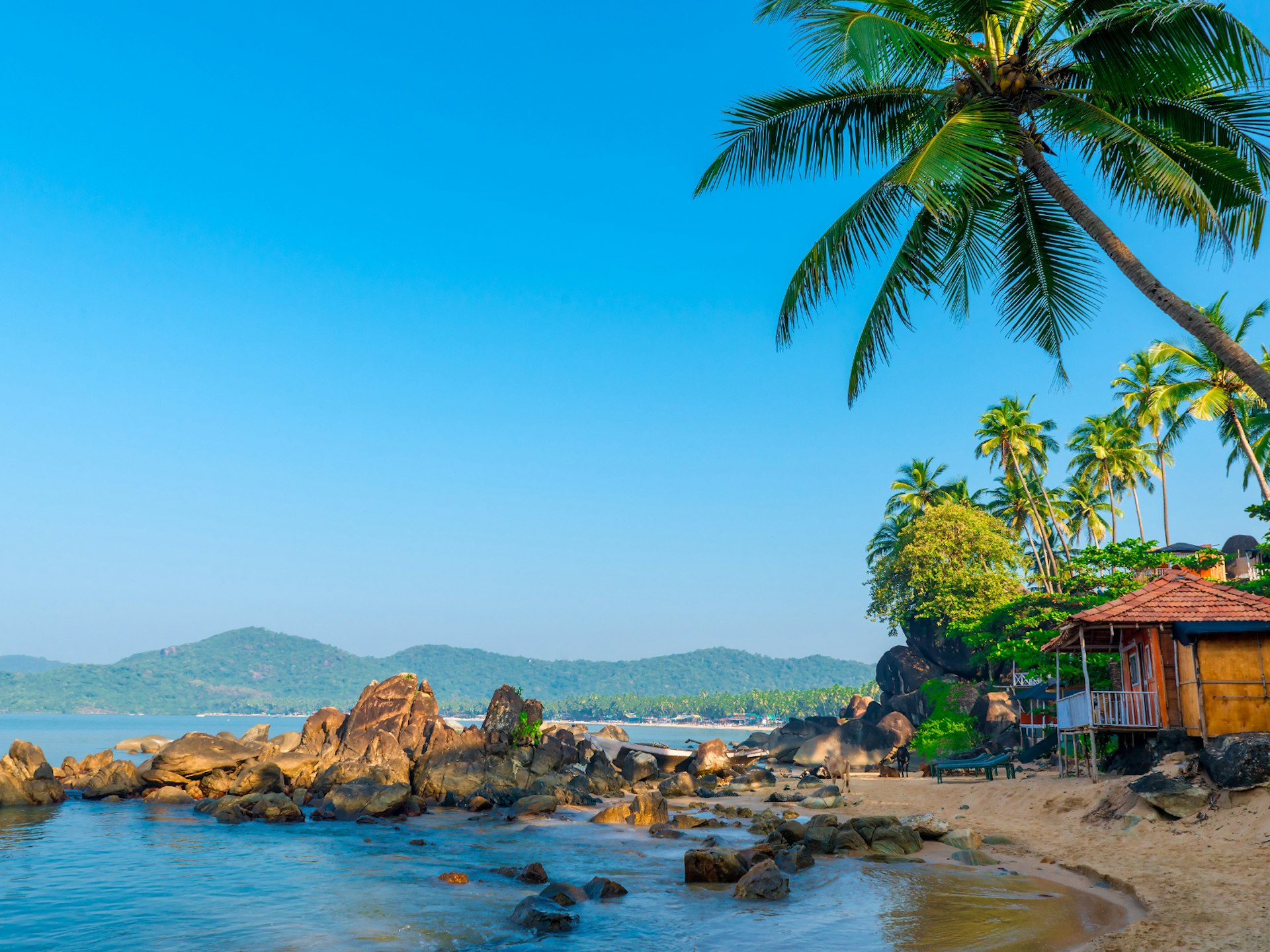A beach in Goa, India with a beach hut, palm trees and a rocky outcrop leading into the ocean