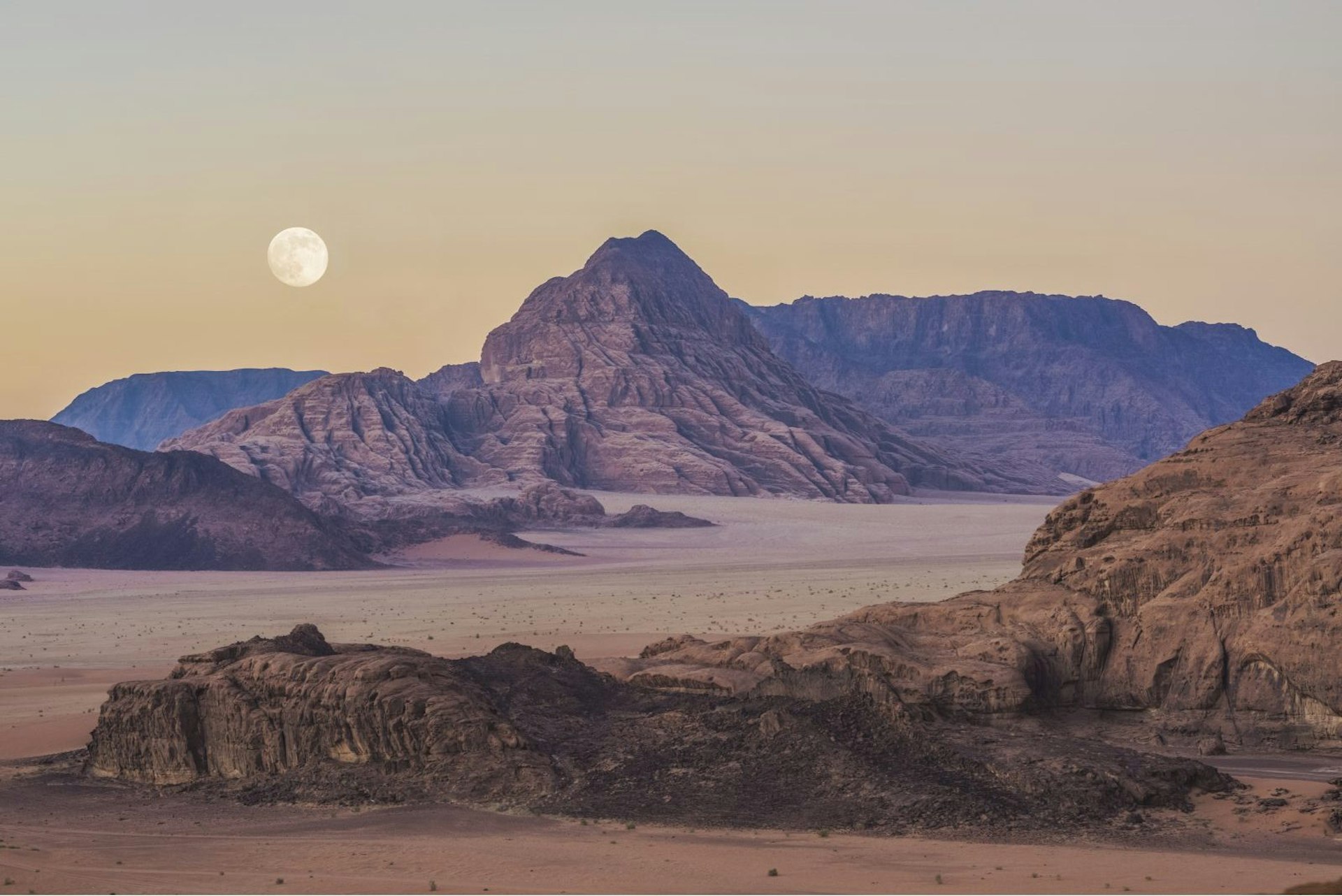 A pale red and brown landscape stretches out, with rocks and hills. The moon can be seen in the sky. The sky is slightly yellow giving the scene an eerie feel