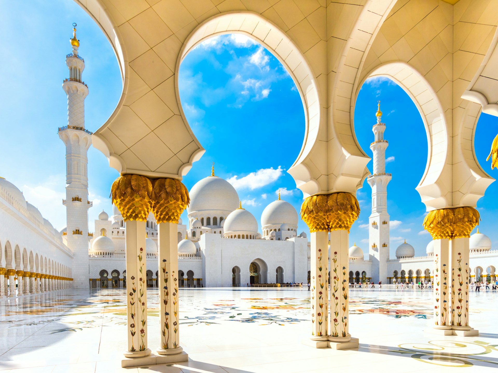 Looking through some archways at the sunlit front of the Sheikh Zayed Mosque with many white domes topped with gold