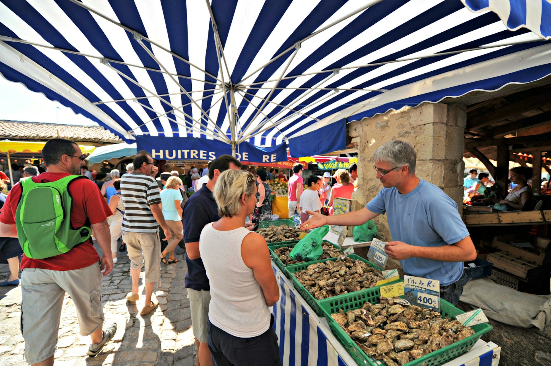 An oyster seller talks with a customer at La Flotte-en-Re market; there is a large blue and white striped awning overhead and other shoppers walk past over the cobbled marketplace.