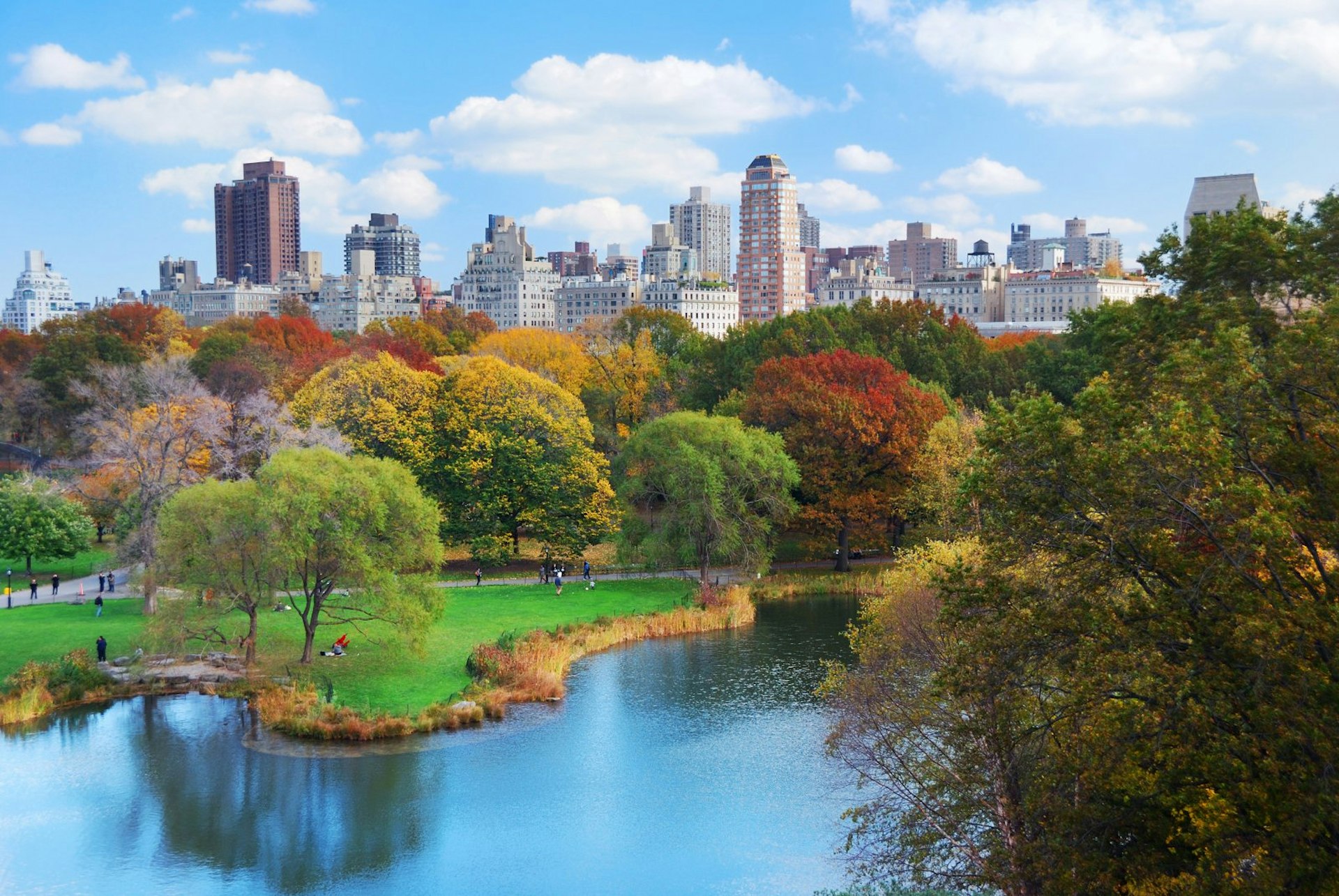 Trees with leaves of yellow, red, brown and green are around a lake in Central Park. Manhattan is seen in the background