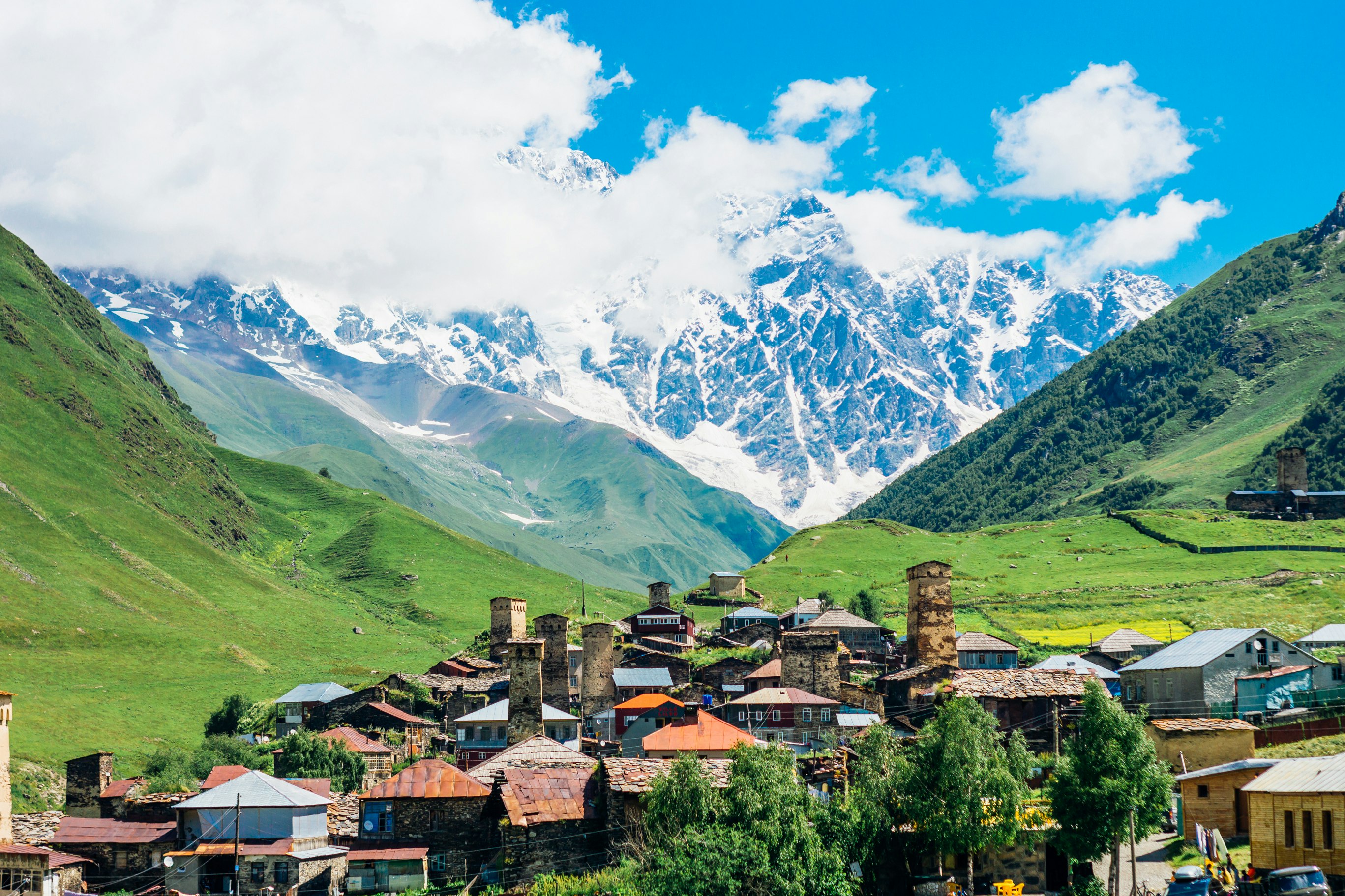 A village with several distinct stone towers standing above the low-rise buildings, in a valley surrounded by large mountains.