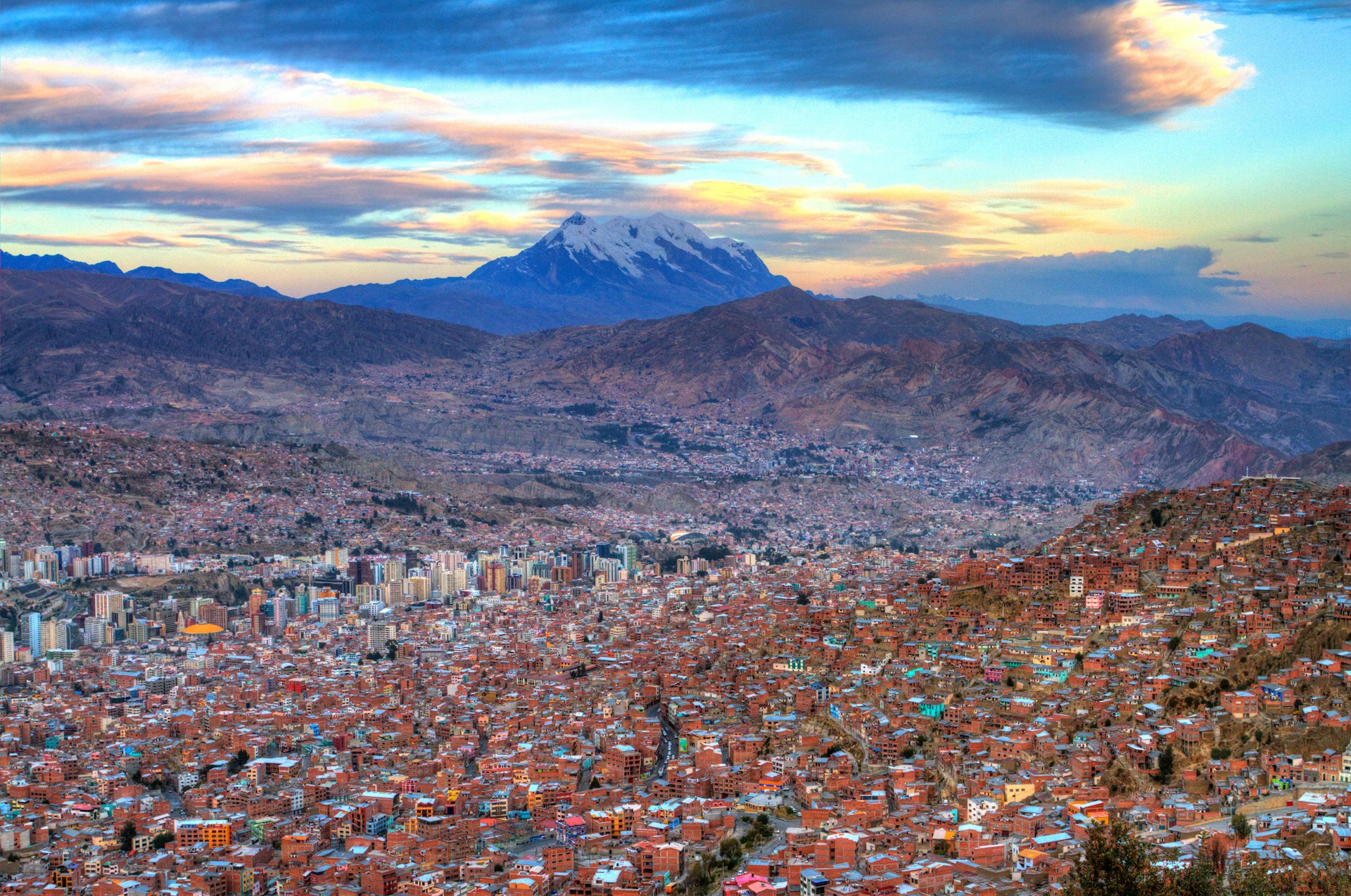 The huge expanse of La Paz, all red buildings and tower blocks, stretching across a high plateau with mountain peaks and a vivid sunset in the distance.