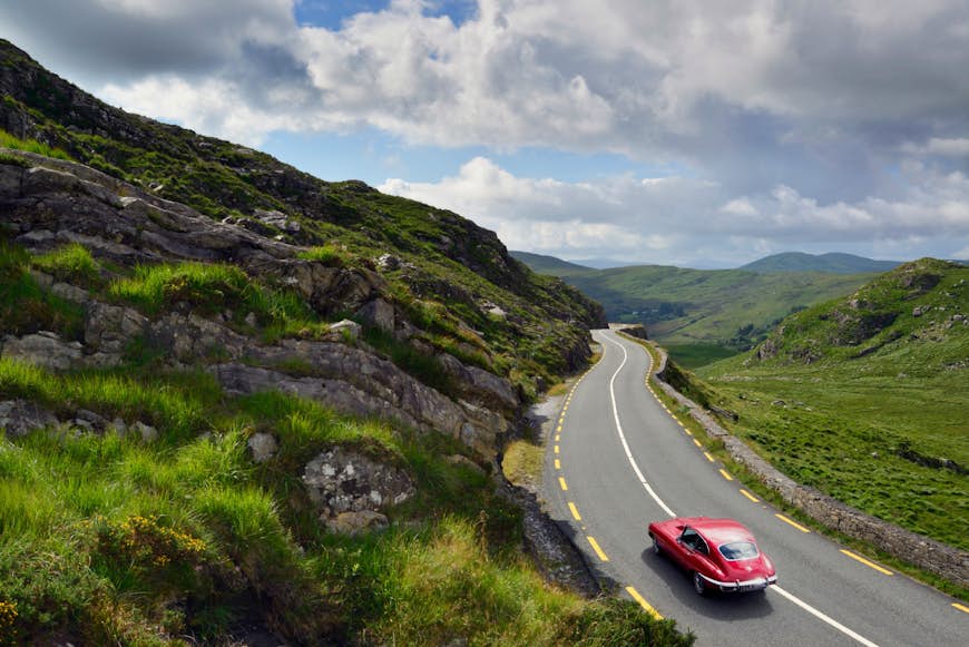 En gammaldags röd bil kör längs en väg som ligger på böljande gröna kullar under en molnig himmel