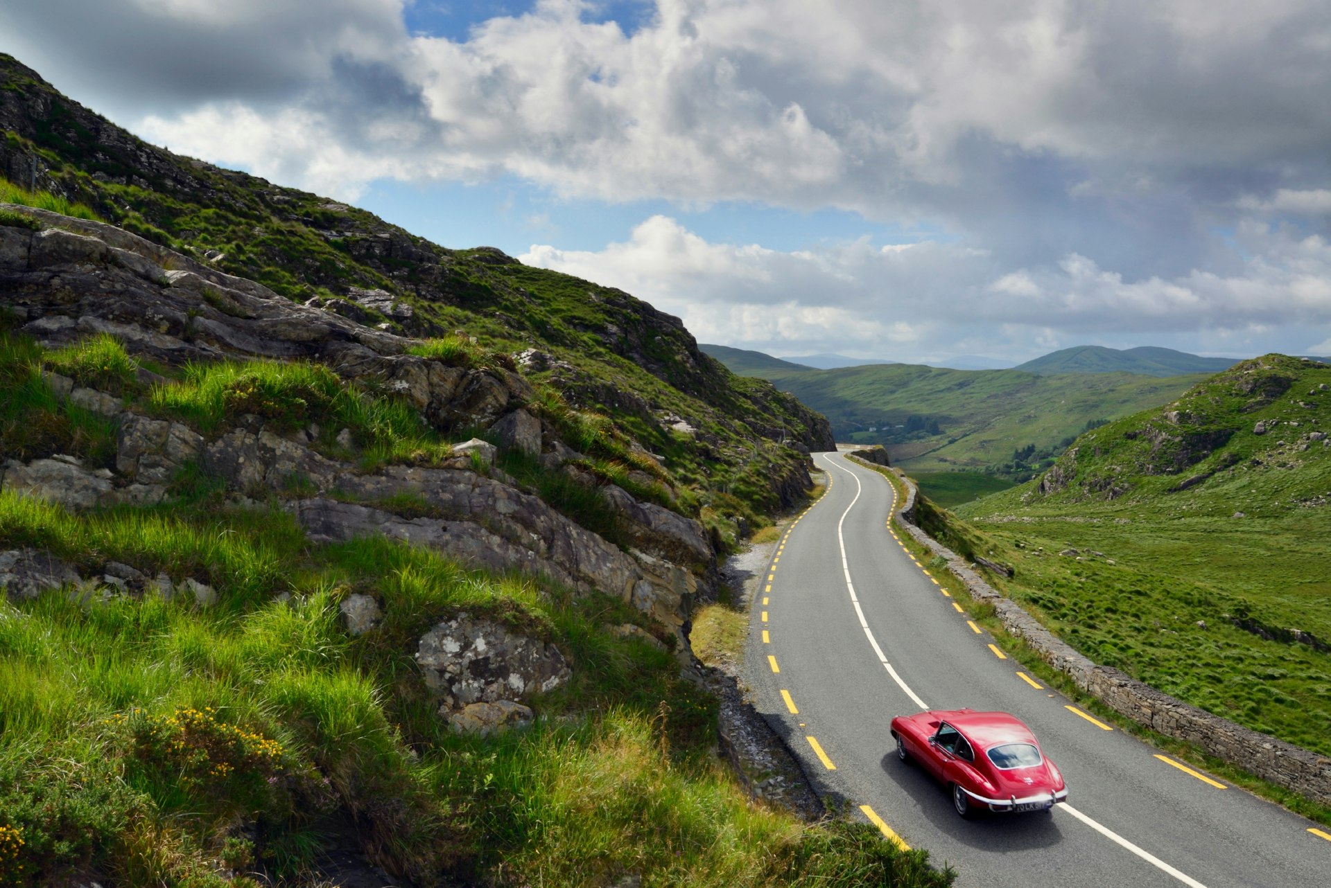 An old-fashioned red car drives along a road set on rolling green hills under a cloudy sky
