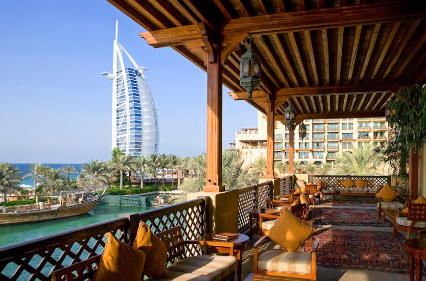 Restaurant with a view of Burj Al Arab in Dubai. The photo is taken from a varnished wooden balcony decorated with woven rugs and wooden, cushioned seats. 