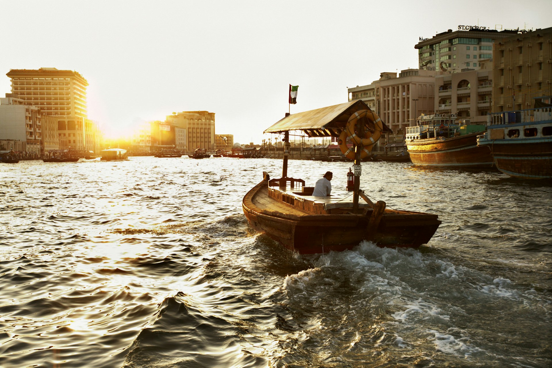 Korsar Dubaiviken på en abra, en traditionell träbåt som används för att taxi människor över bäcken.  Solen står lågt på skyline och delvis skymd av kontorsbyggnader.  En ensam man styr båten och det finns inga passagerare. 
